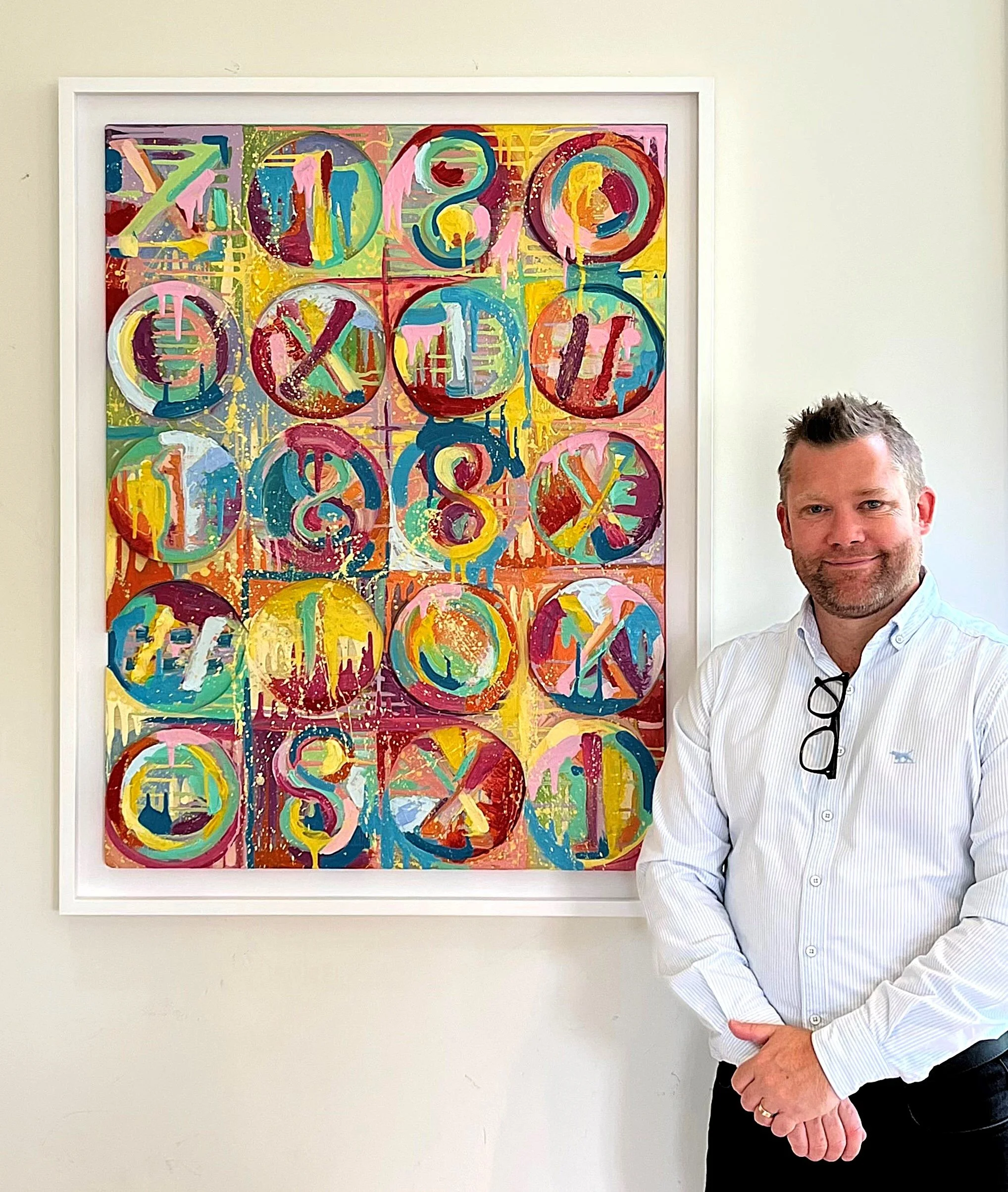 A man with glasses hanging from his shirt stands next to a colorful abstract painting featuring painted numbers and circles.