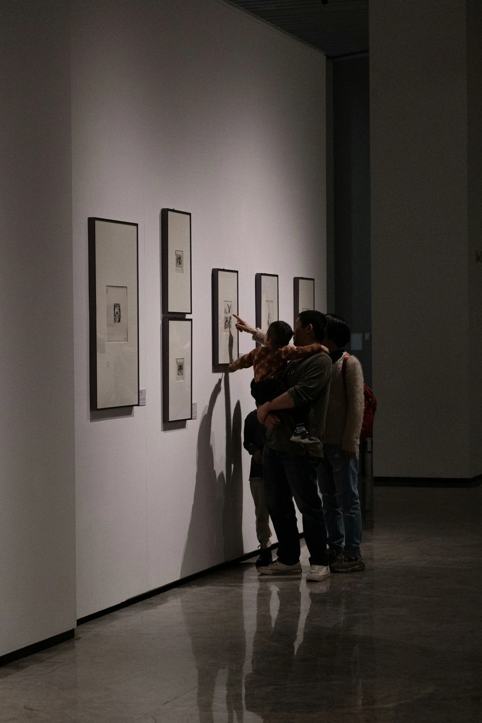 Three people, including a child, looking at artwork on a white wall in a gallery, with the child pointing at one piece.