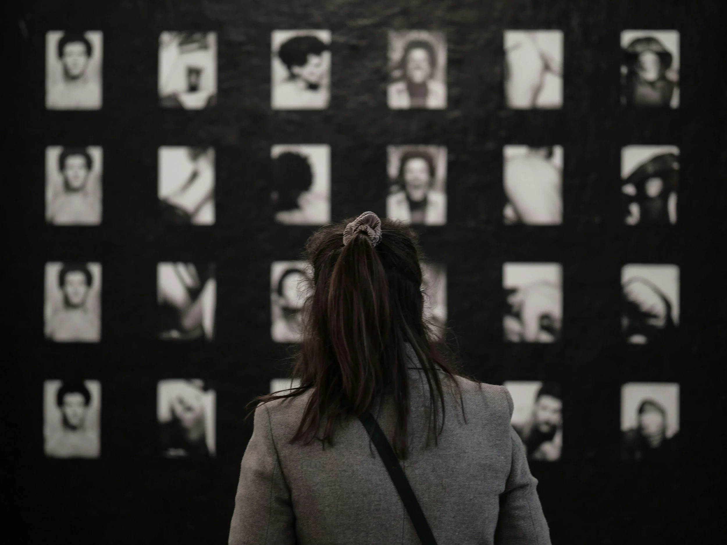 A woman with dark hair tied in a ponytail watching black and white portrait photographs on a black wall.