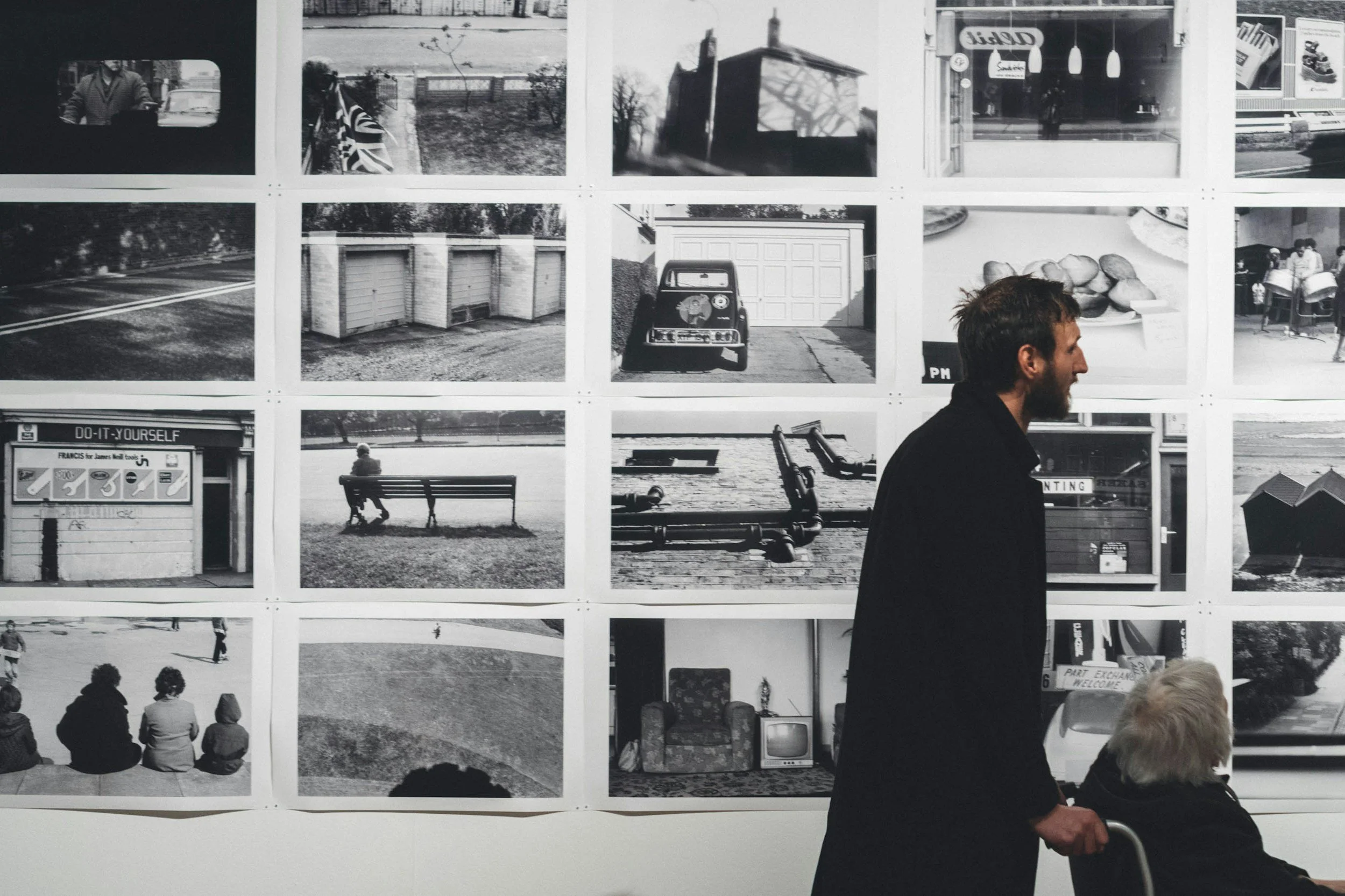 Black and white photograph of a man and an elderly woman with a walker in front of a wall displaying a grid of black and white photographs of a loved one who has died.