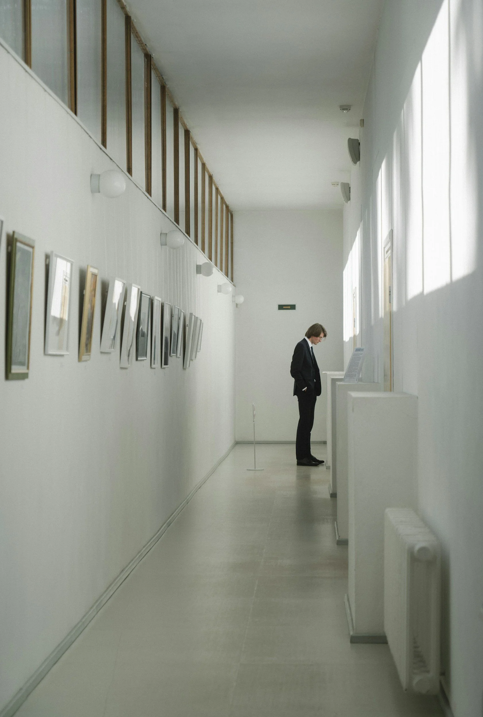 A person in a black suit and glasses looking at photos of a loved one in a gallery corridor with white walls and a row of pictures on the left, natural light coming through windows on the right.