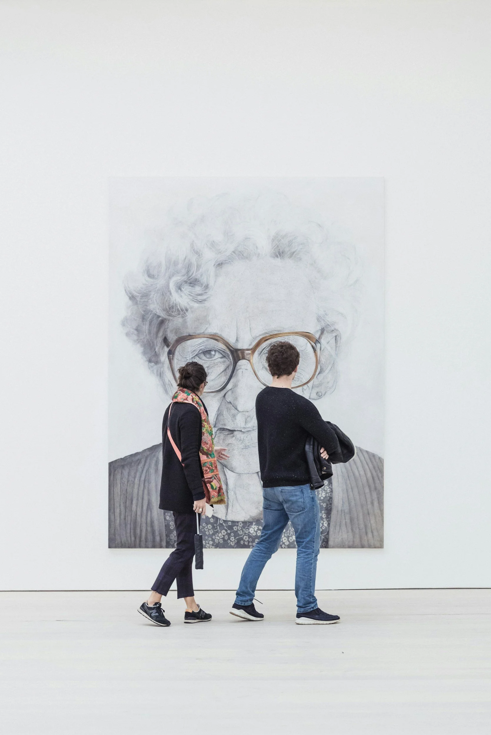 Two people walking past a large black-and-white portrait of an elderly woman with curly hair, wearing glasses and a patterned shirt, displayed on a white wall in an art gallery.