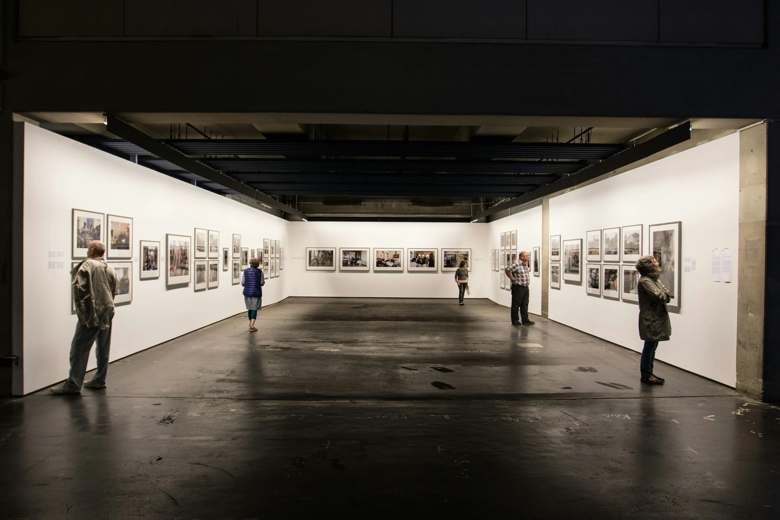 People viewing framed photographs in an art gallery with white walls and black ceiling beams.