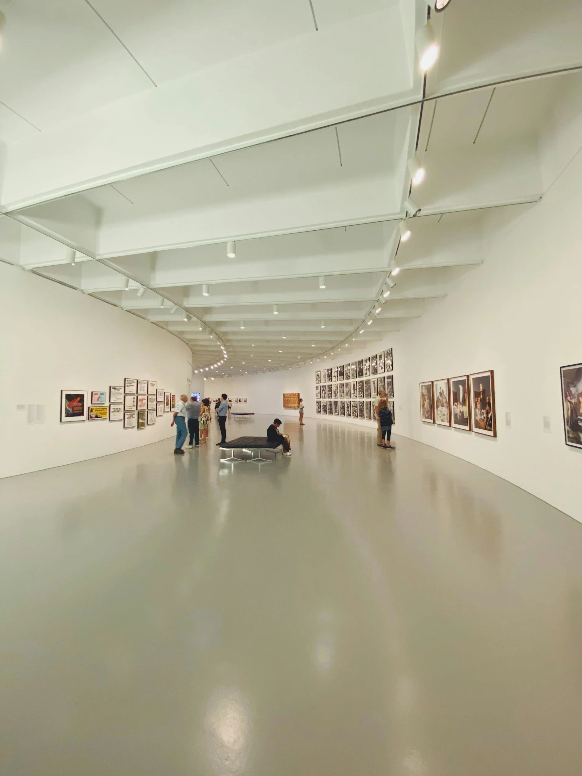 An art gallery with white walls displaying framed artwork and photographs. Visitors are walking around and observing the exhibits. There is a sitting area with a black bench in the center of the gallery.