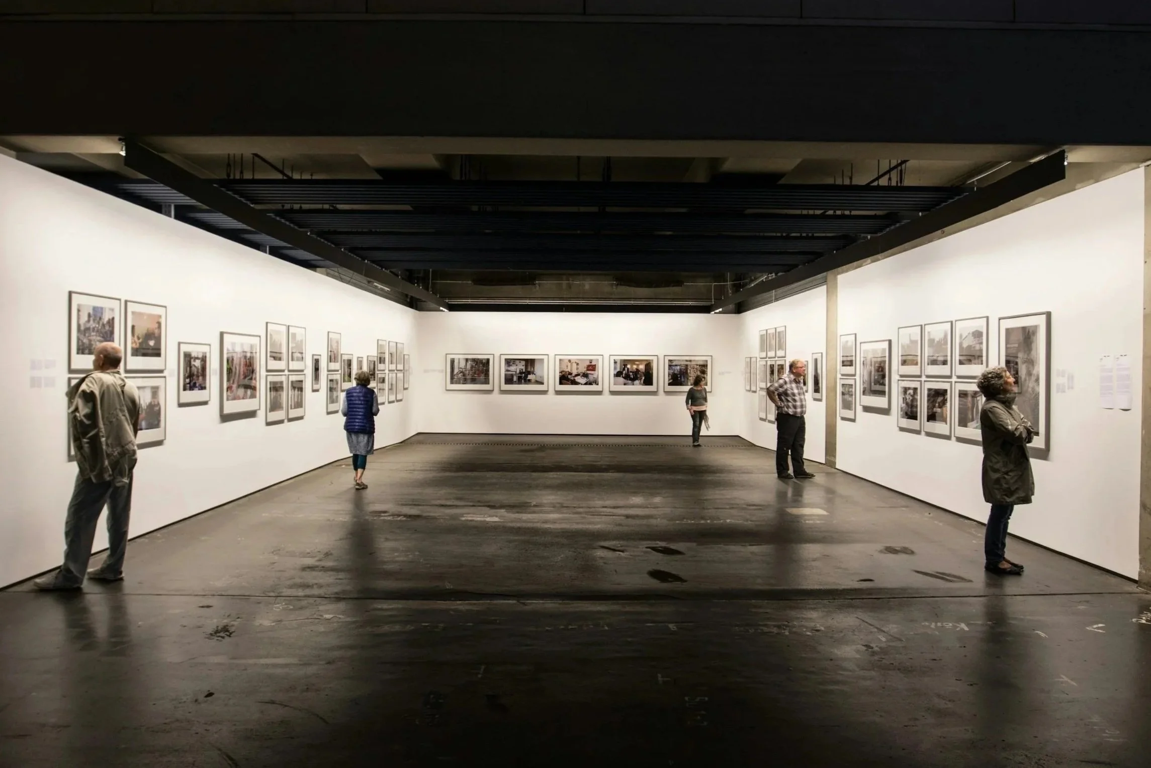 People viewing framed photographs displayed on white gallery walls in an art exhibition gallery with wooden floors.