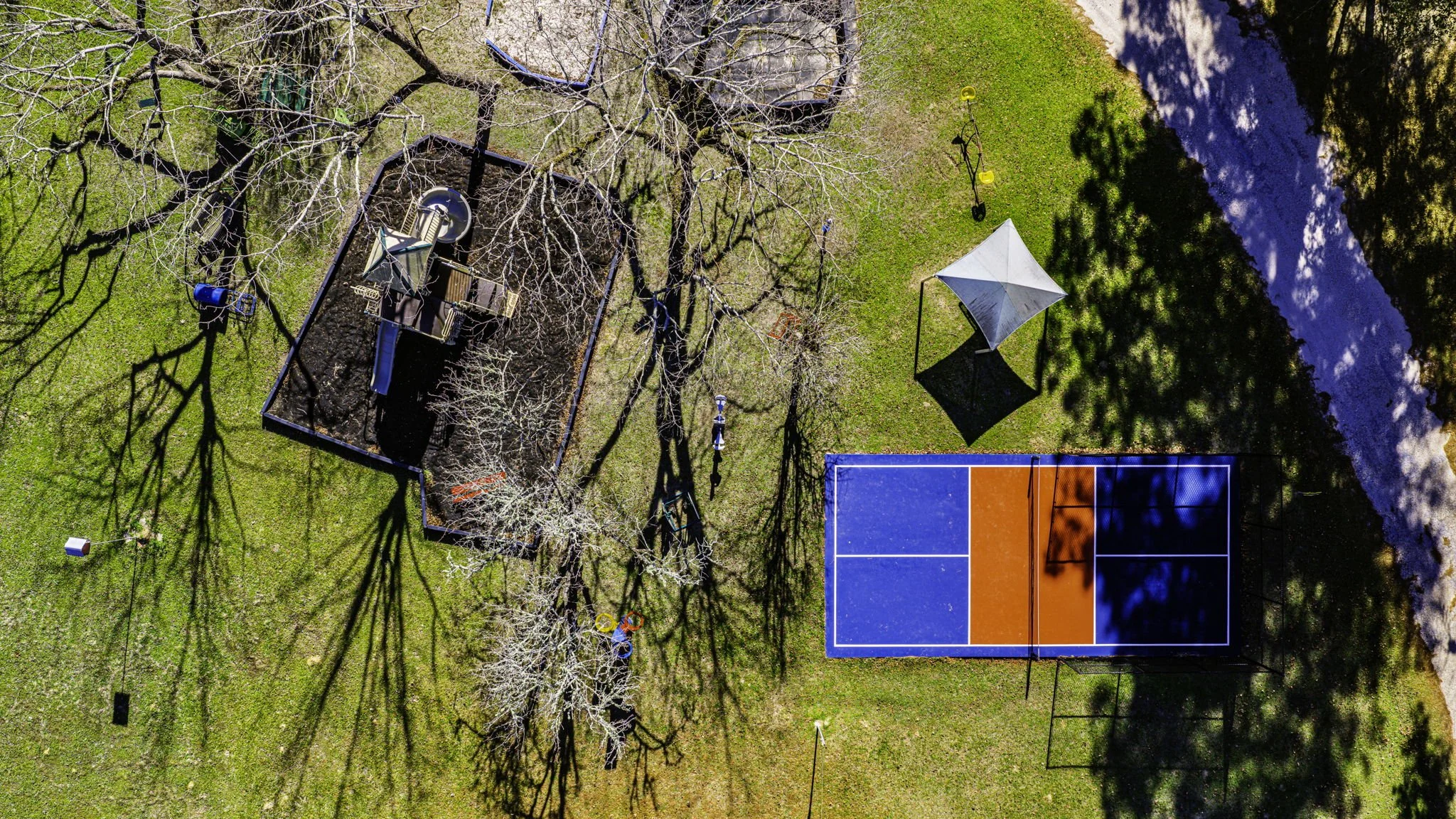 An aerial view of a playground with a sandbox, swings, and a slide surrounded by trees, a shaded sitting area, a basketball court, and a walking pathway.