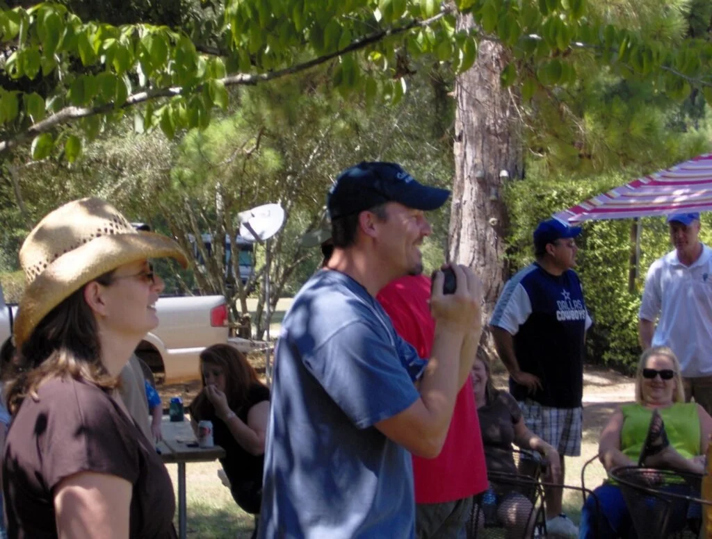 Group of people outdoors in a shaded park, smiling and chatting, with some seated and others standing, under trees and with a picnic table.