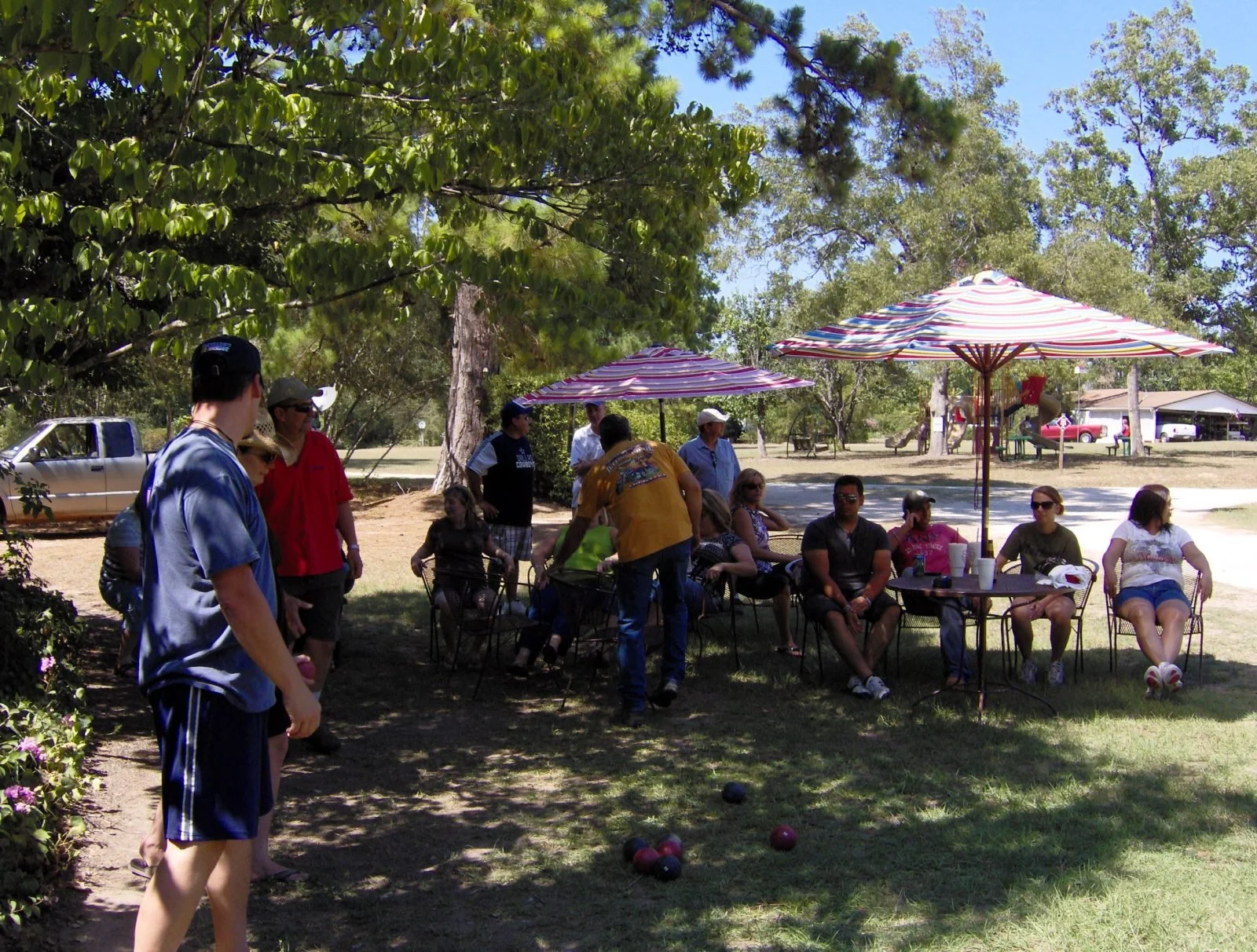 Group of people gathered outdoors under trees, some sitting at tables with striped umbrellas, others standing, with a playground and picnic area in the background.