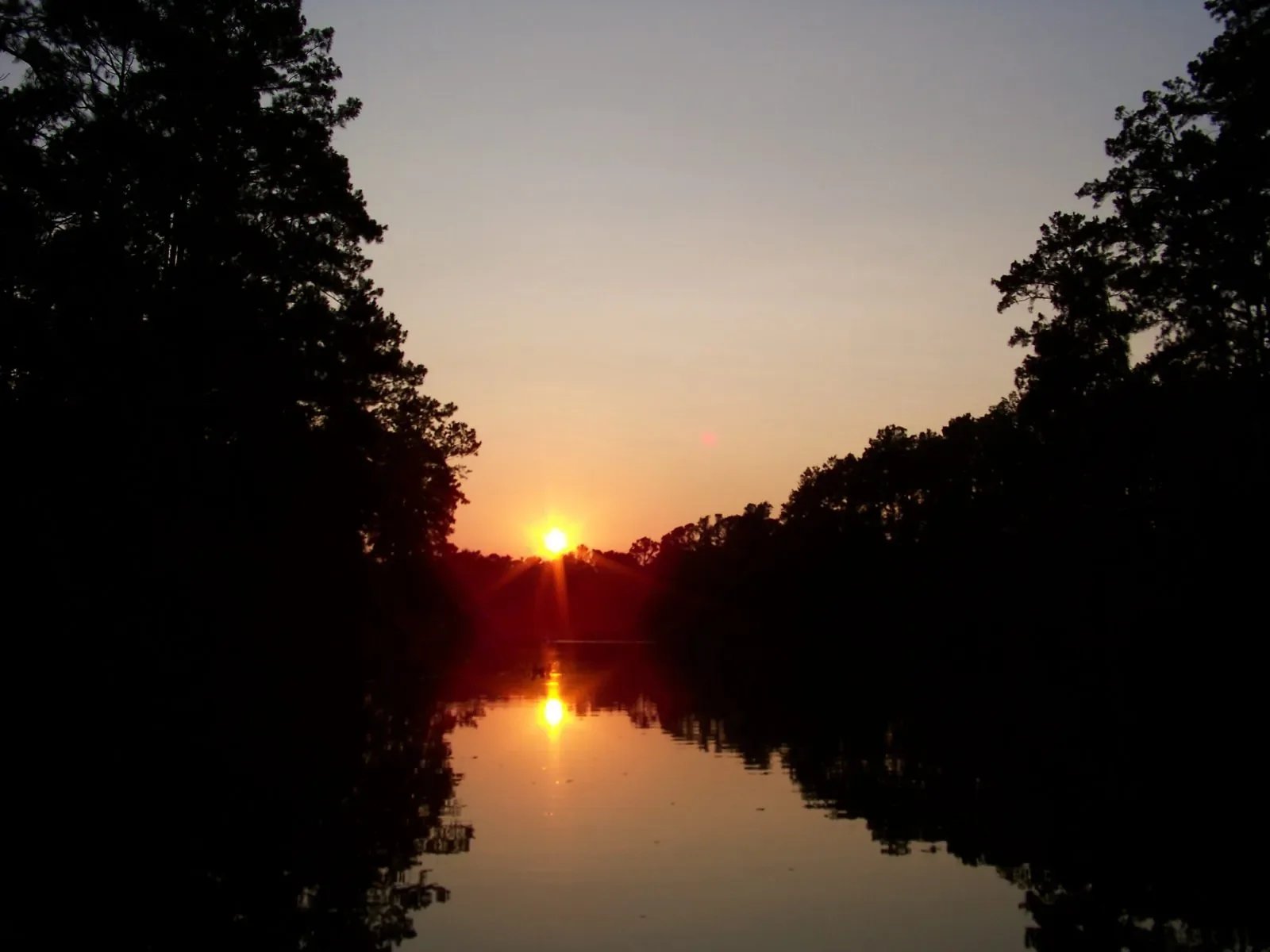 Sunset over a calm river surrounded by dark silhouettes of trees, with the sun low on the horizon, casting a reflection on the water.