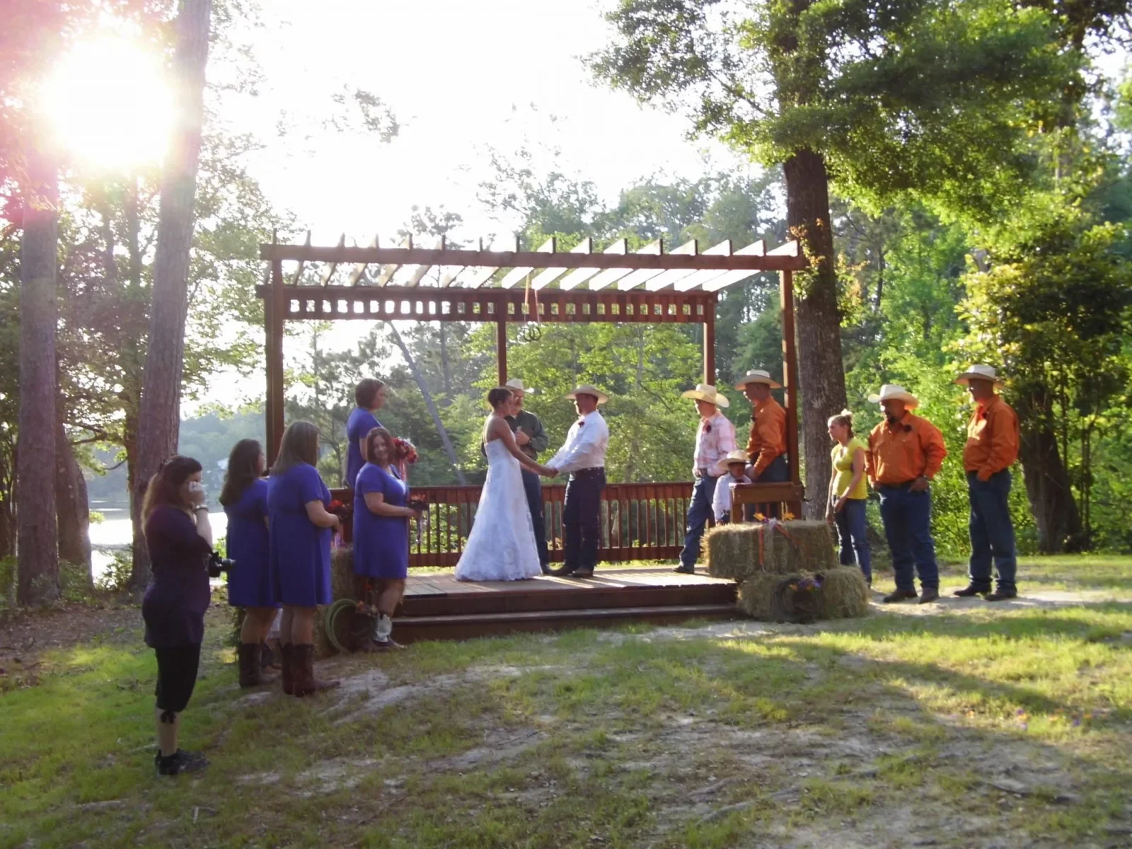 A wedding ceremony outdoors on a wooden platform surrounded by trees and sunlight, with a bride and groom exchanging rings, and guests standing nearby.