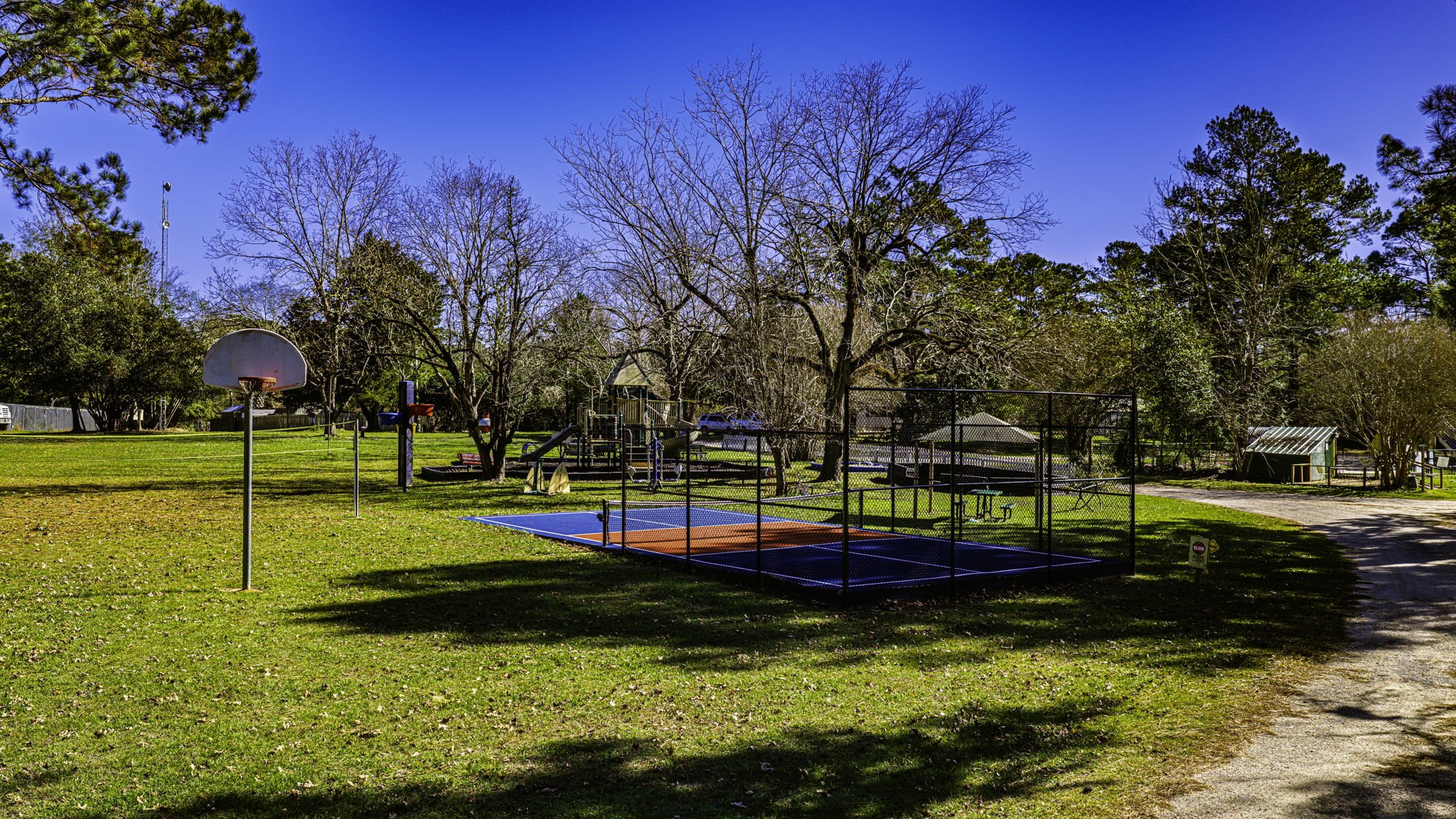 A park with a basketball hoop, a fenced tennis or pickleball court, a picnic area with benches and tables, a small shed, and trees under a bright blue sky.