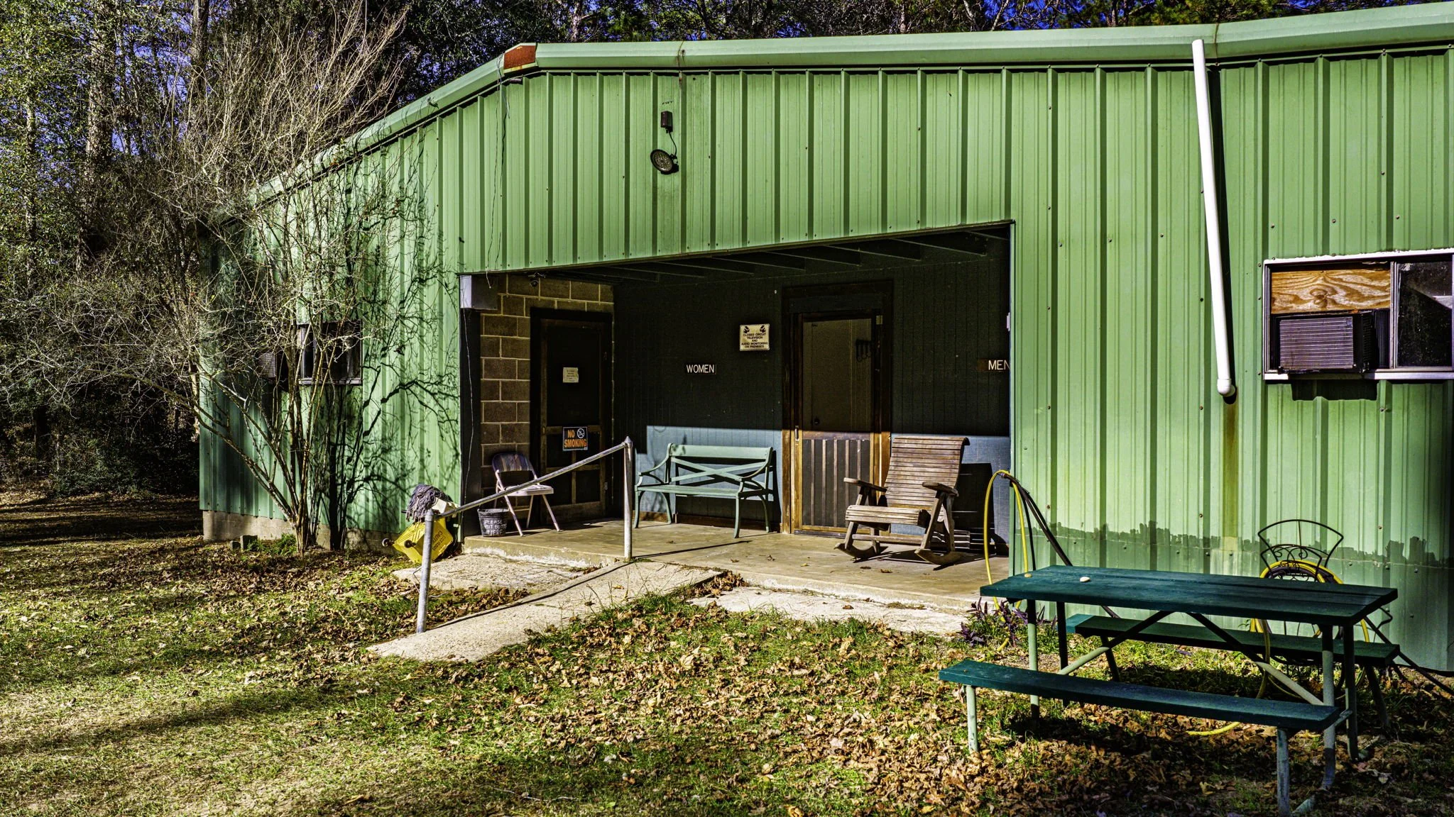 Green outdoor restroom building with separate men's and women's entrances, surrounded by trees and leaves, outdoor chairs and tables, and a concrete walkway.