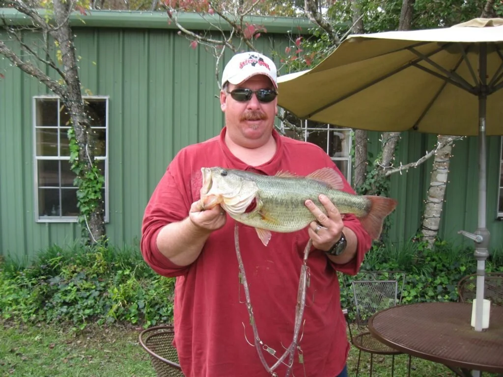 A man wearing sunglasses, a baseball cap, and a red shirt is holding a large fish with both hands. The fish appears to be a bass. The background shows a green building, trees, a patio umbrella, and outdoor furniture.