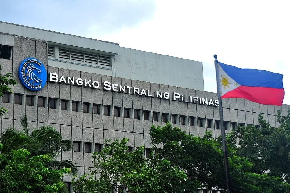 A large gray government building with the sign 'Bangko Sentral ng Pilipinas,' the central bank of the Philippines. A Filipino flag is flying in front of the building, with green trees partially obscuring the lower part of the structure.
