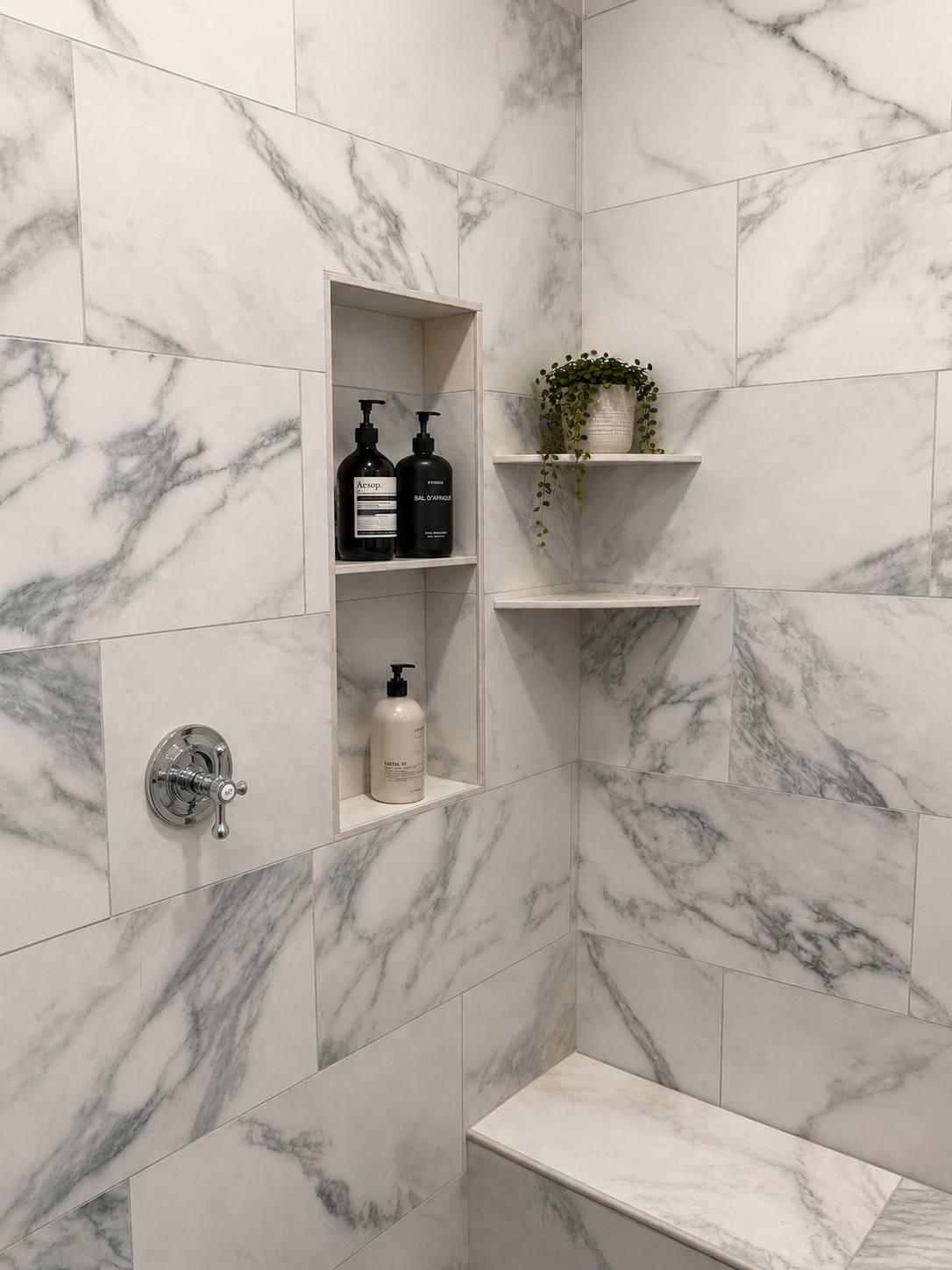 Marble tiled shower with built-in niche holding shampoo and soap bottles, and corner shelves with a potted plant above a marble bench.
