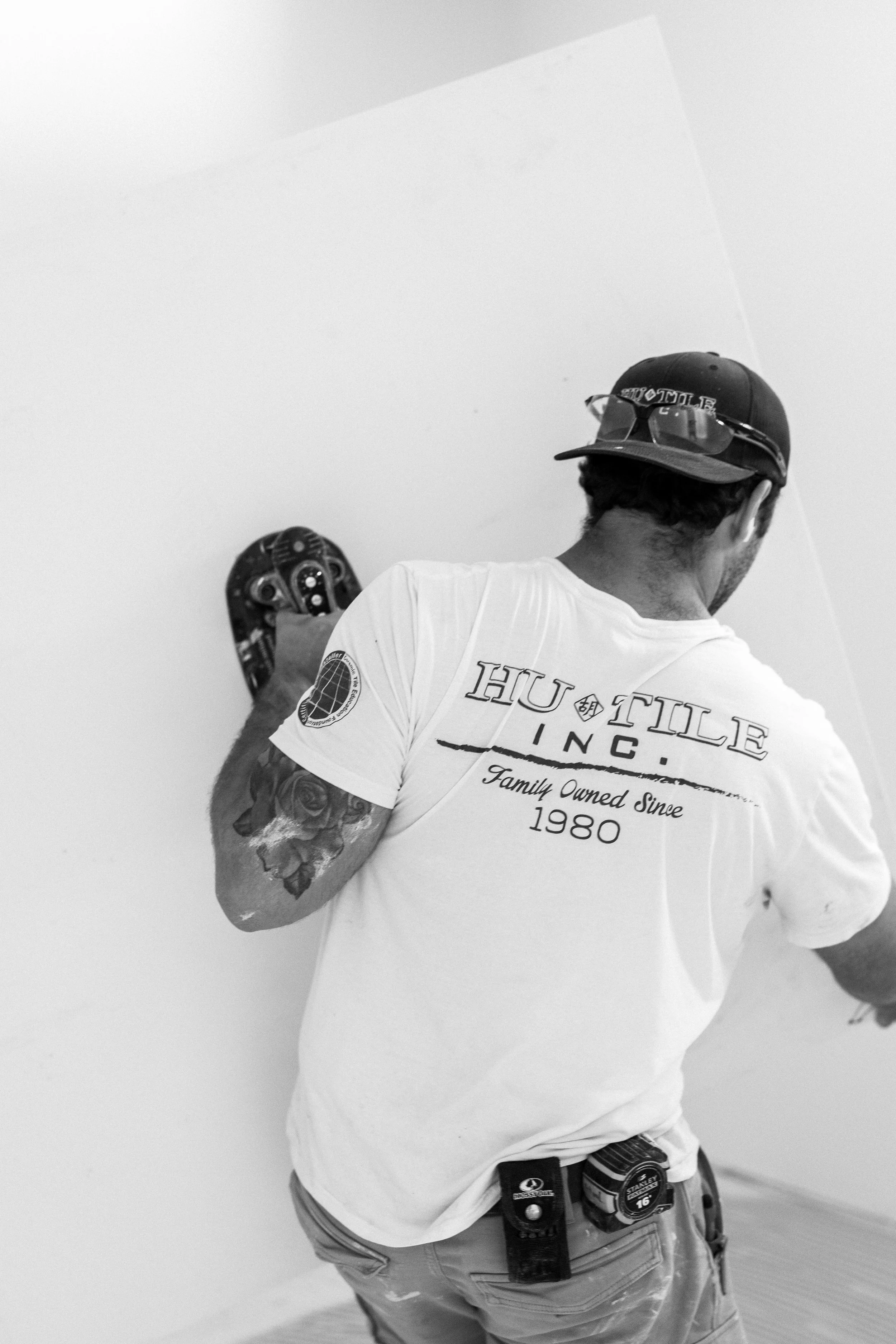 A man with tattoos on his arm near the shoulder wearing a white T-shirt with the text "HUT TUL INCl. Family Owned Since 1980," and a black cap with sunglasses resting on top. He is standing indoors in front of a plain, light-colored wall, with construction tools tucked into his belt, including a power drill. The image is in black and white.