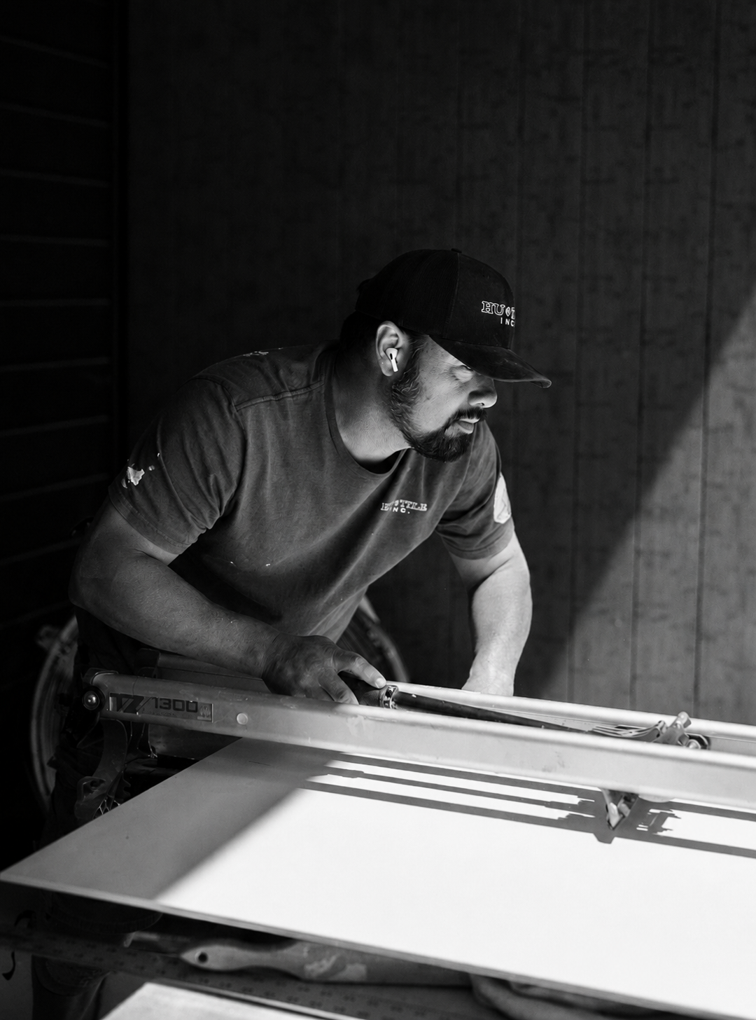 A man working on a large flat surface, possibly a table or cutting board, in a dimly lit room. He is wearing a cap, t-shirt, and wireless earbuds.