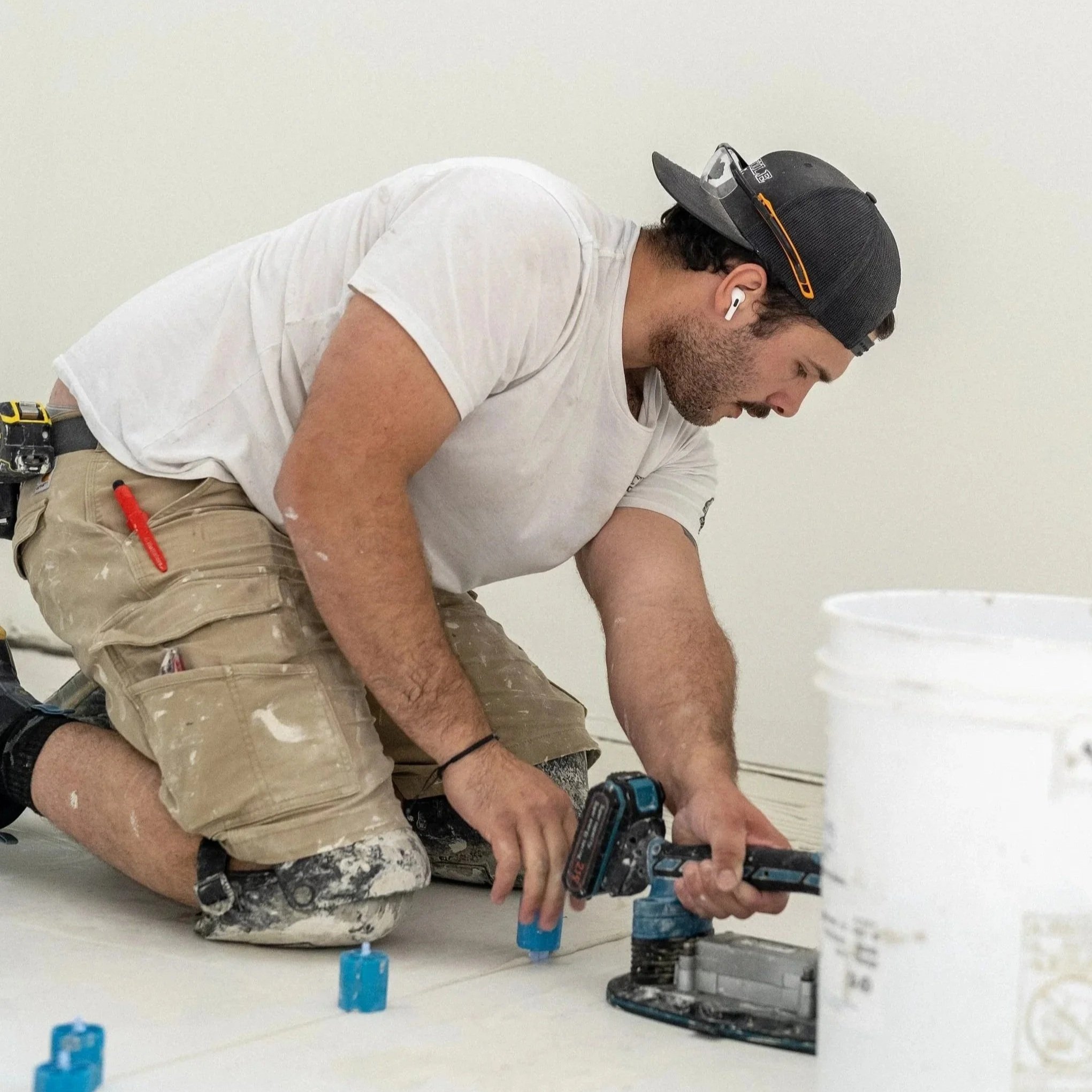 A man working on a construction project, kneeling on the floor, using power tools, wearing a cap, white t-shirt, beige shorts with paint splatters, and ear protection.
