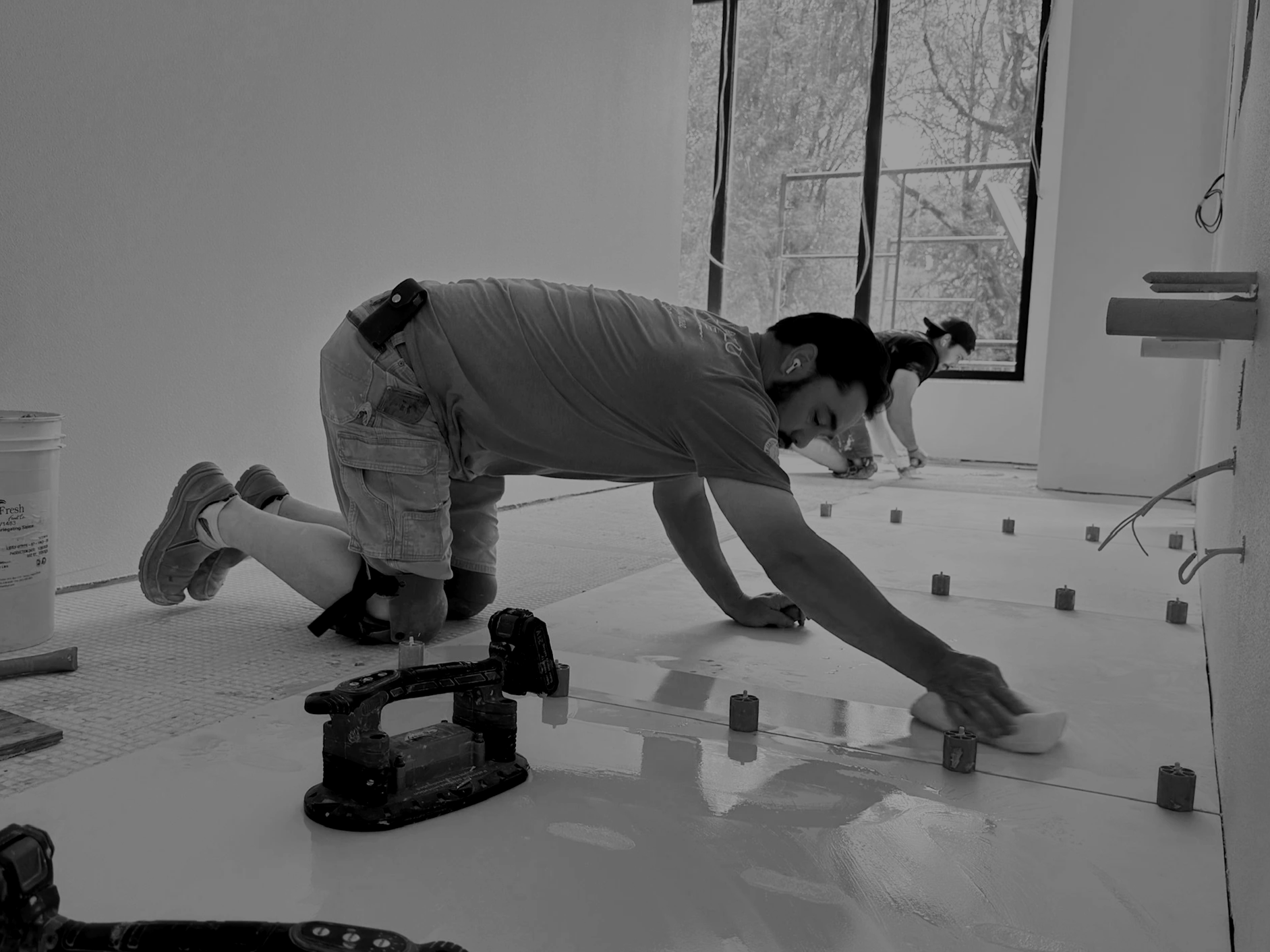 Construction workers installing a floor, with one kneeling and smoothing surface with a trowel, near tools, along with a woman working in the background.