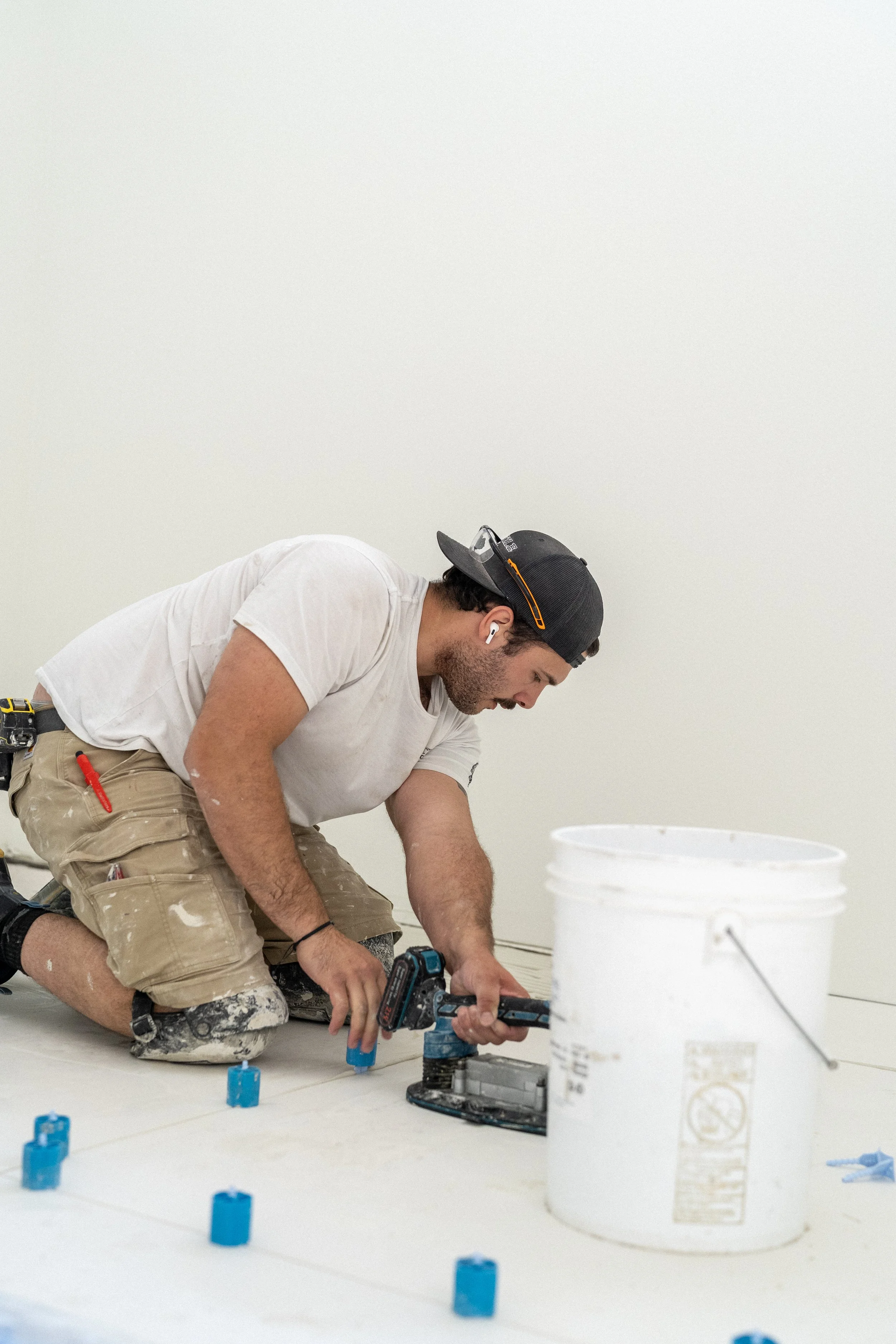 A man wearing a white t-shirt, khaki shorts, a black cap, and white earbud headphones is kneeling on the floor, working with a power drill. There are blue plastic spacers on the floor around him. A large white bucket is in the foreground, and he appears to be installing or working on flooring or wall tiles.