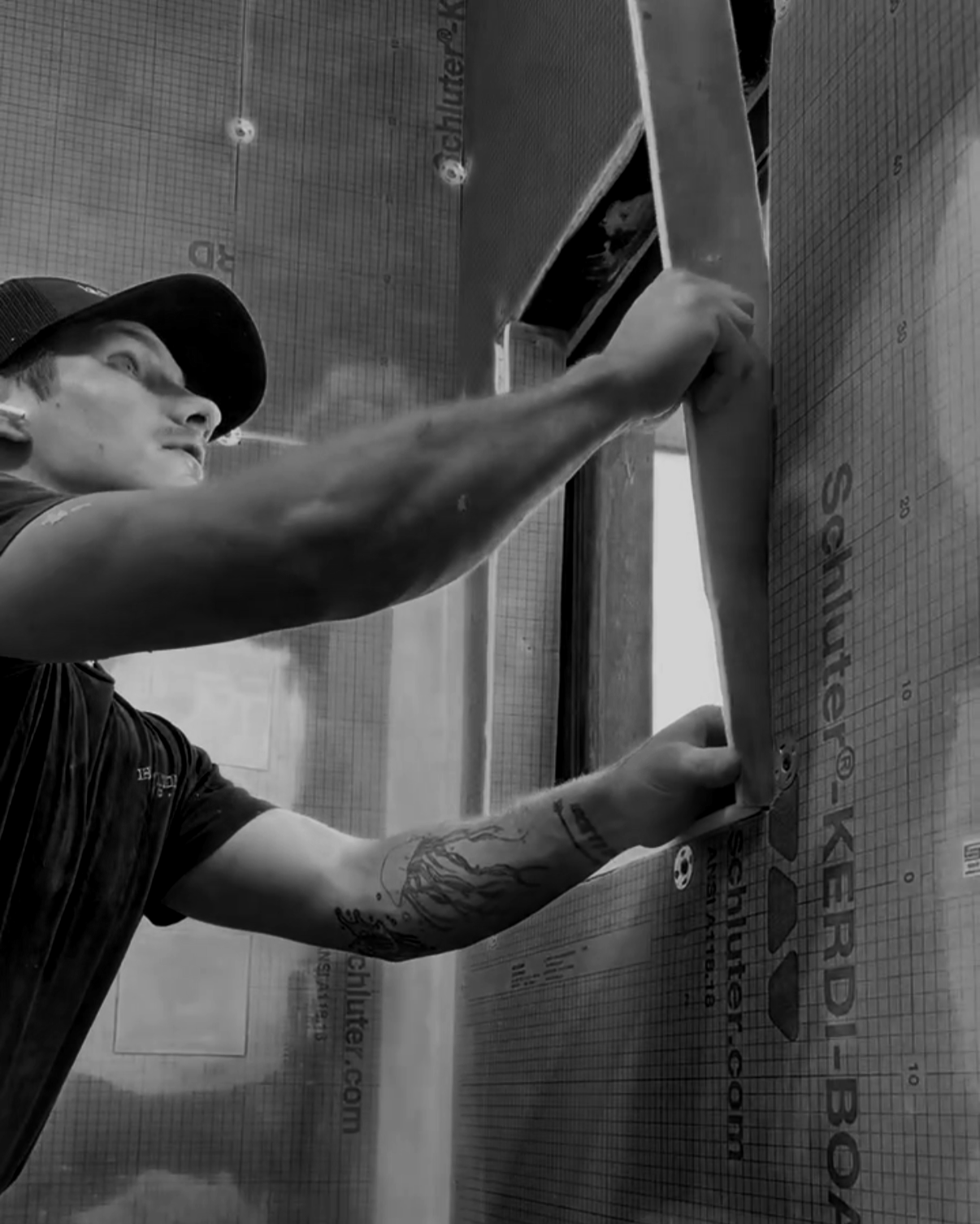 A person installing or repairing a window, wearing a black cap and T-shirt, tattooed arm, in an indoor setting with a wall covered in labels and a grid pattern.