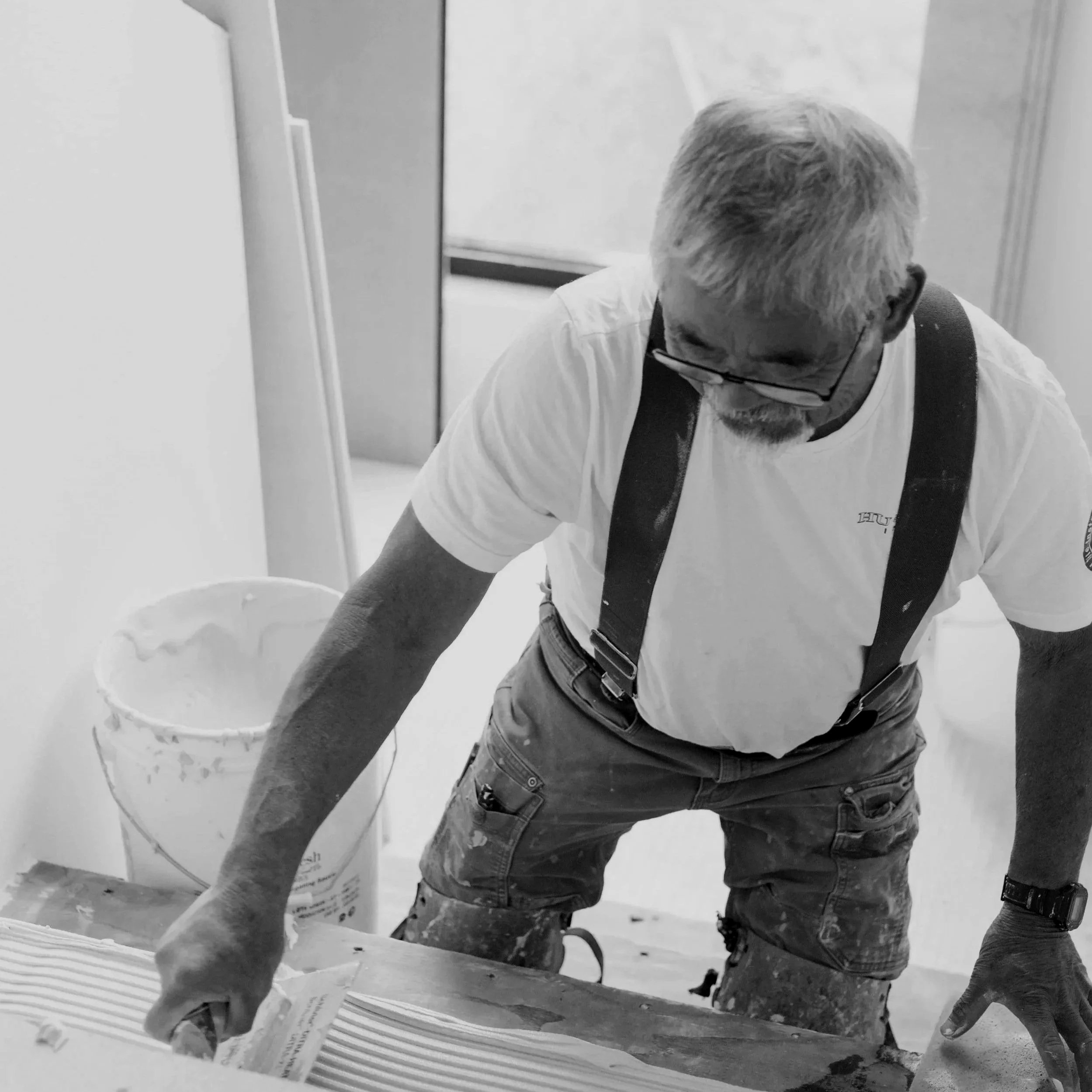 Two workers are installing or setting up a piece of industrial equipment or structure in a workshop or construction area. One worker is wearing glasses, a white shirt, and a backpack, leaning over with hands on the equipment. The other worker, wearing a dark jacket, is also leaning over and assisting. A large container or bag is on the floor nearby, and various equipment and materials are scattered around.