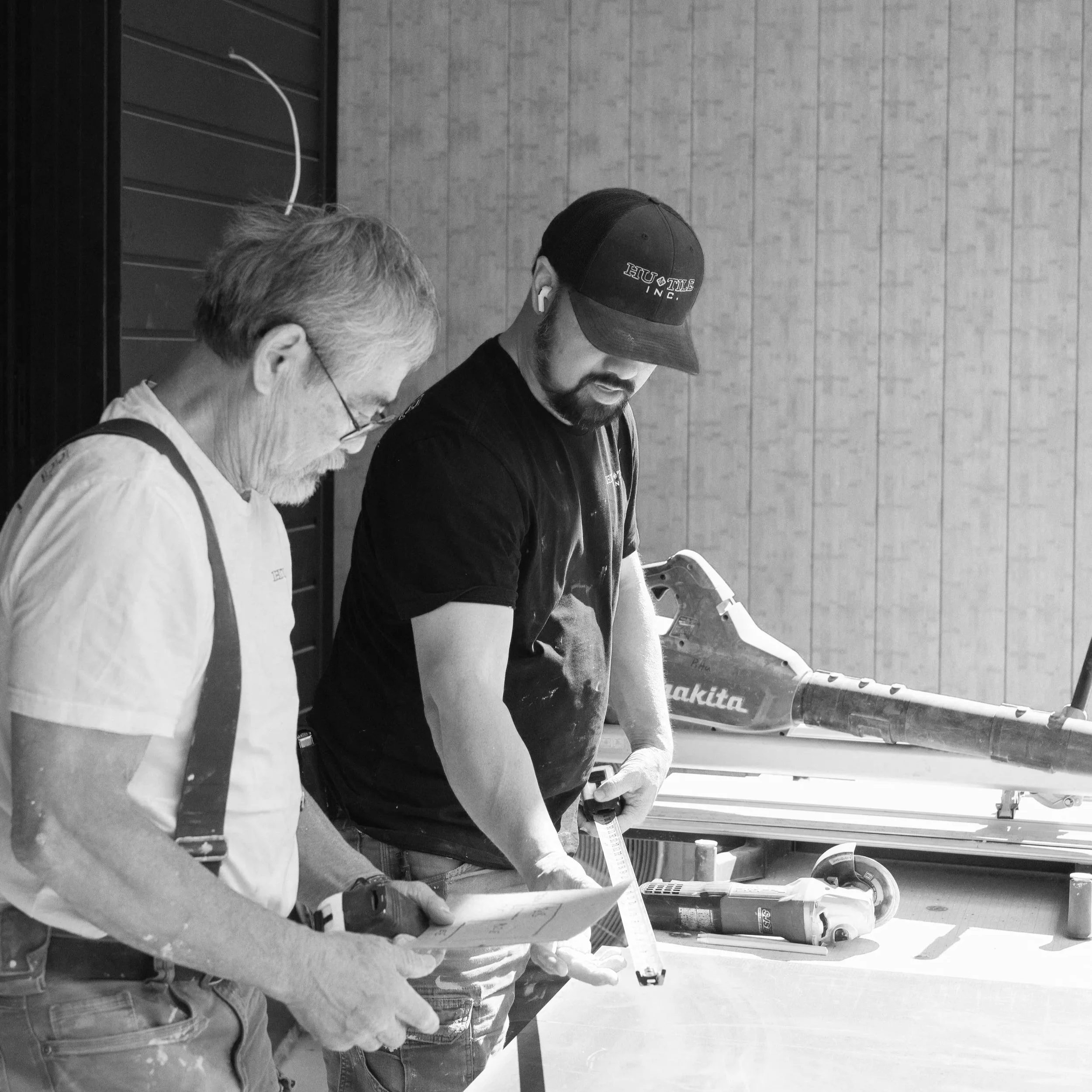 Two men are working together in a workshop, reviewing plans or documents with various tools and equipment around them.