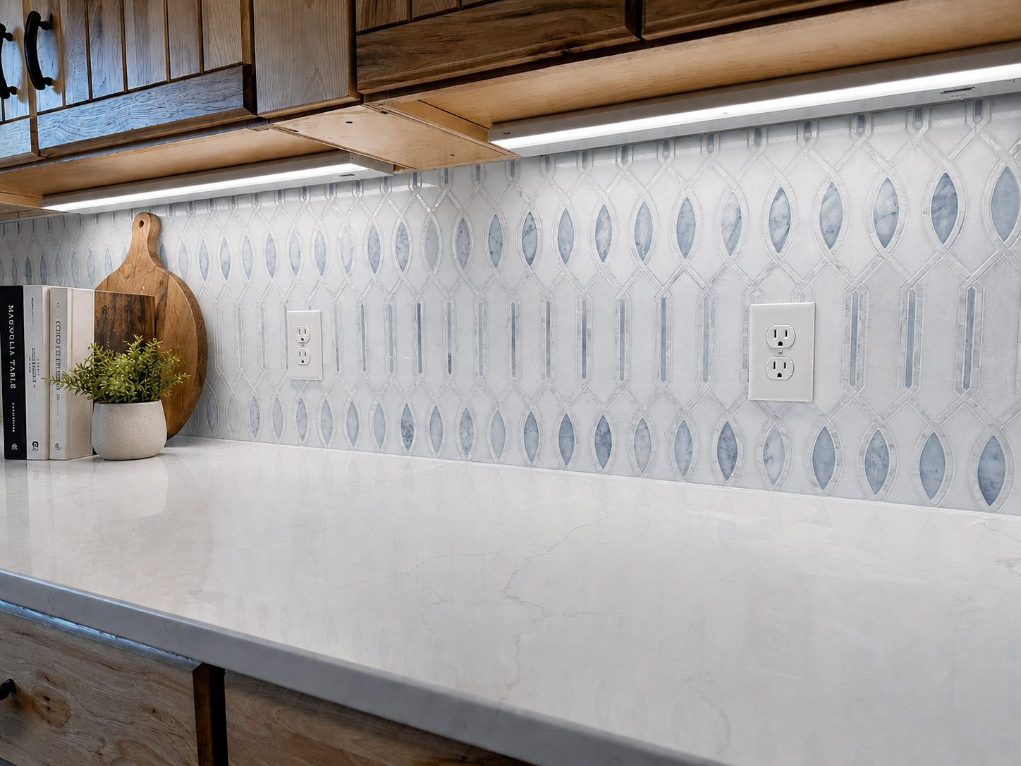 Kitchen counter with white marble surface, decorative tile backsplash with geometric pattern, two electrical outlets, a wooden cutting board, a small potted plant, and some stacked books.