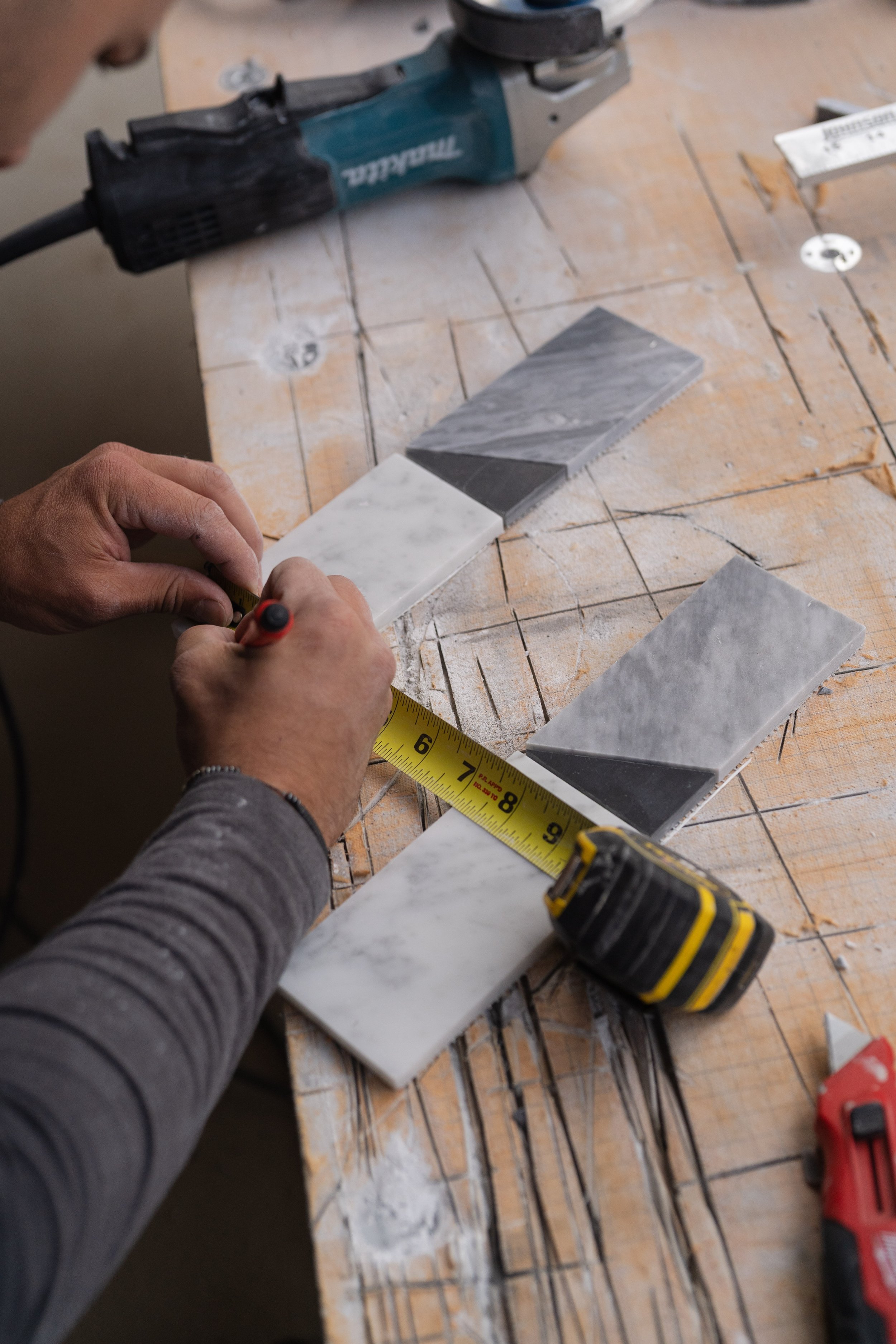 Person measuring and marking marble tiles on a wooden workbench with tools including a tile saw, tape measure, and drill.
