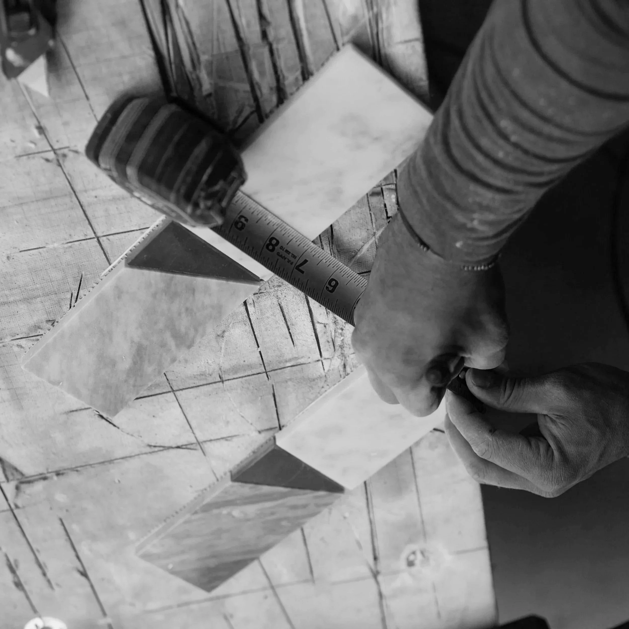 A person's hand holding a ruler next to a small wooden block on a tiled surface, with a piece of paper underneath.