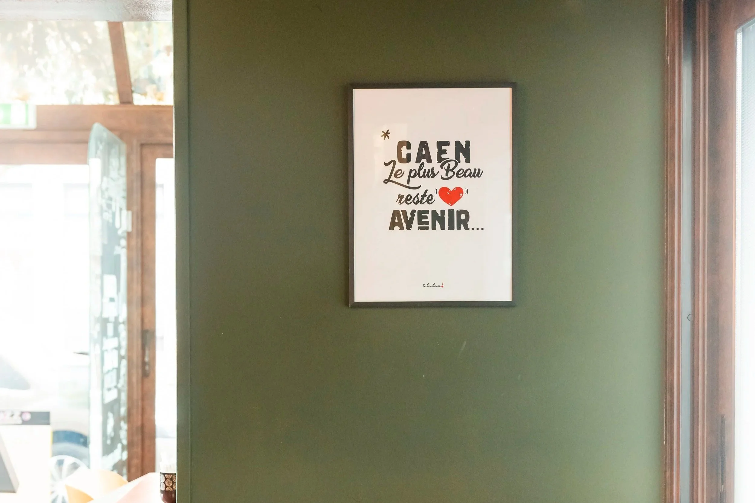 Une affiche avec une citation en français sur un mur vert. La citation dit : "Caen le plus beau reste à venir" avec un cœur rouge illustré.