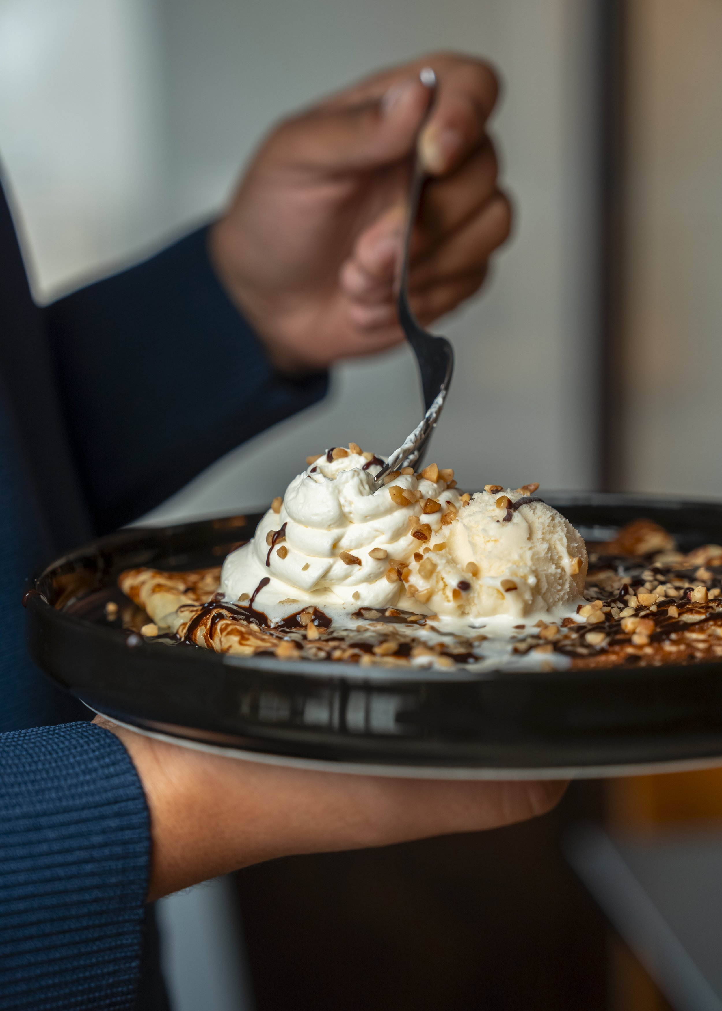 Un dessert avec de la crème glacée, de la crème fouettée, du chocolat, des noix concassées sur une assiette noire, tenu par une personne avec une main visible.