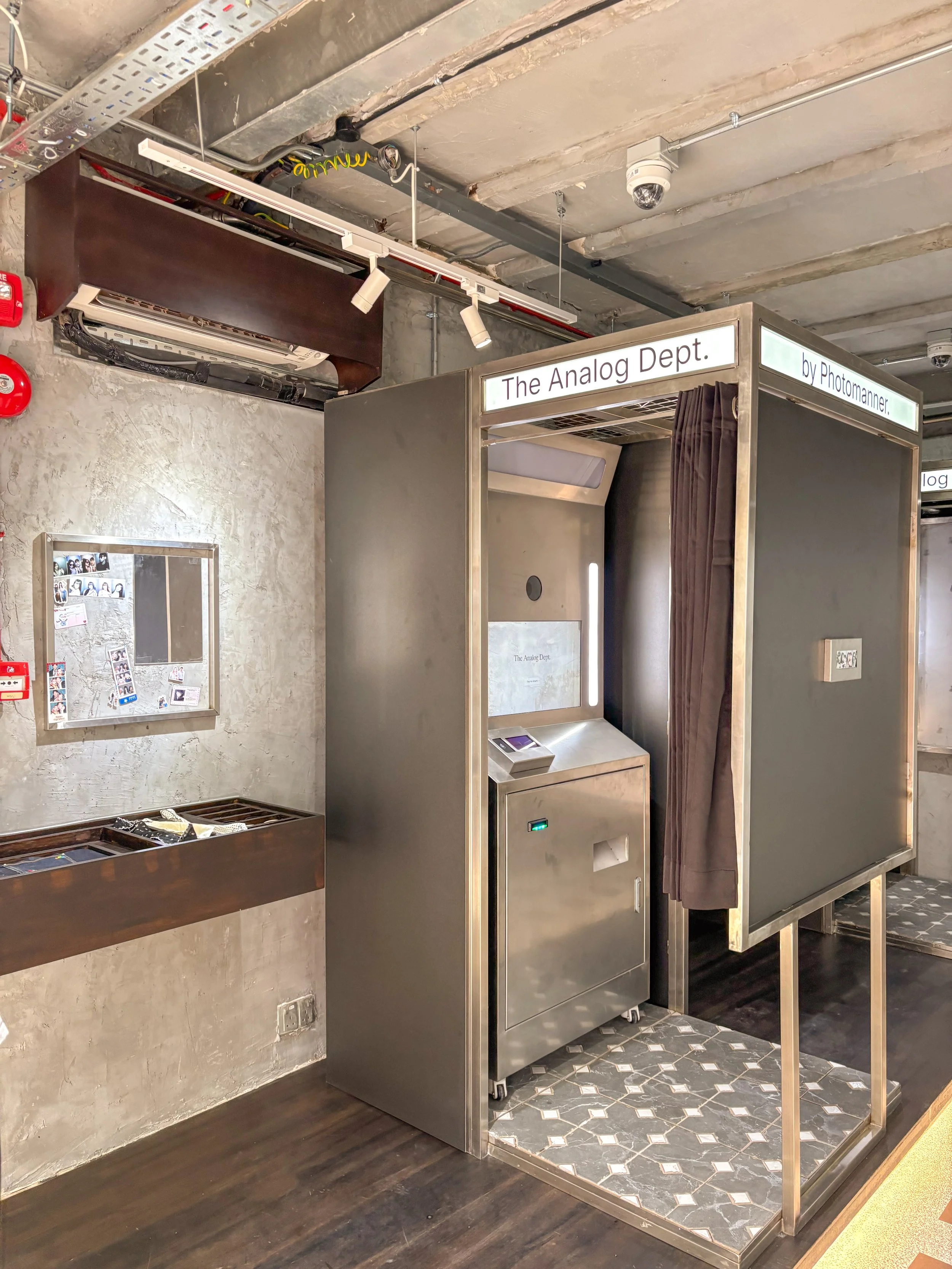 Photo of a photo booth labeled 'The Analog Dept. by Photomanner' in an indoor space with concrete walls and wood flooring. The booth has dark gray and metallic exterior with a privacy curtain and a small screen and printer on the side.