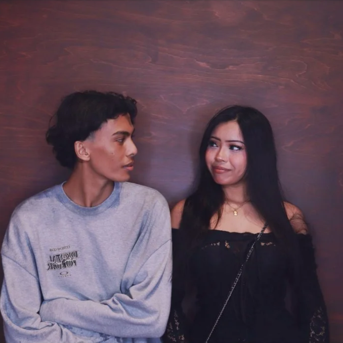 A young man and woman standing together against a wooden wall, looking at each other. The man has curly dark hair and is wearing a gray sweatshirt. The woman has long dark hair, is dressed in black, and is wearing a gold necklace.