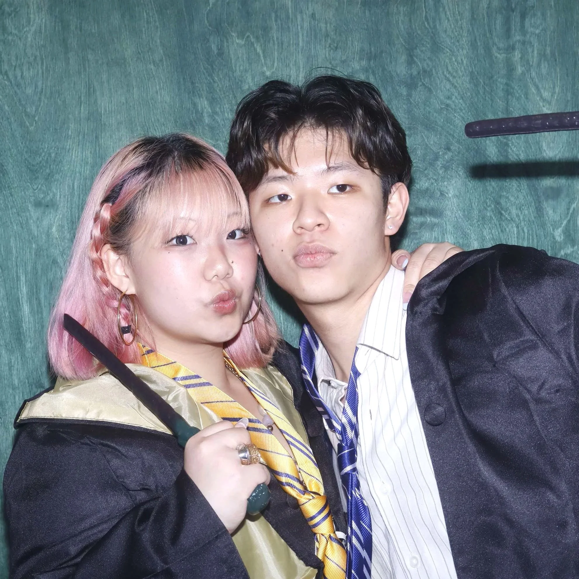 Close-up of two people, a woman with pink hair and a man with dark hair, taking a selfie together. They are dressed in stylish clothing with a green textured background.
