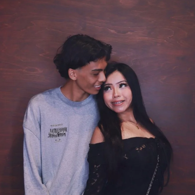 A young man and woman standing close together against a wooden background, smiling and looking at each other.