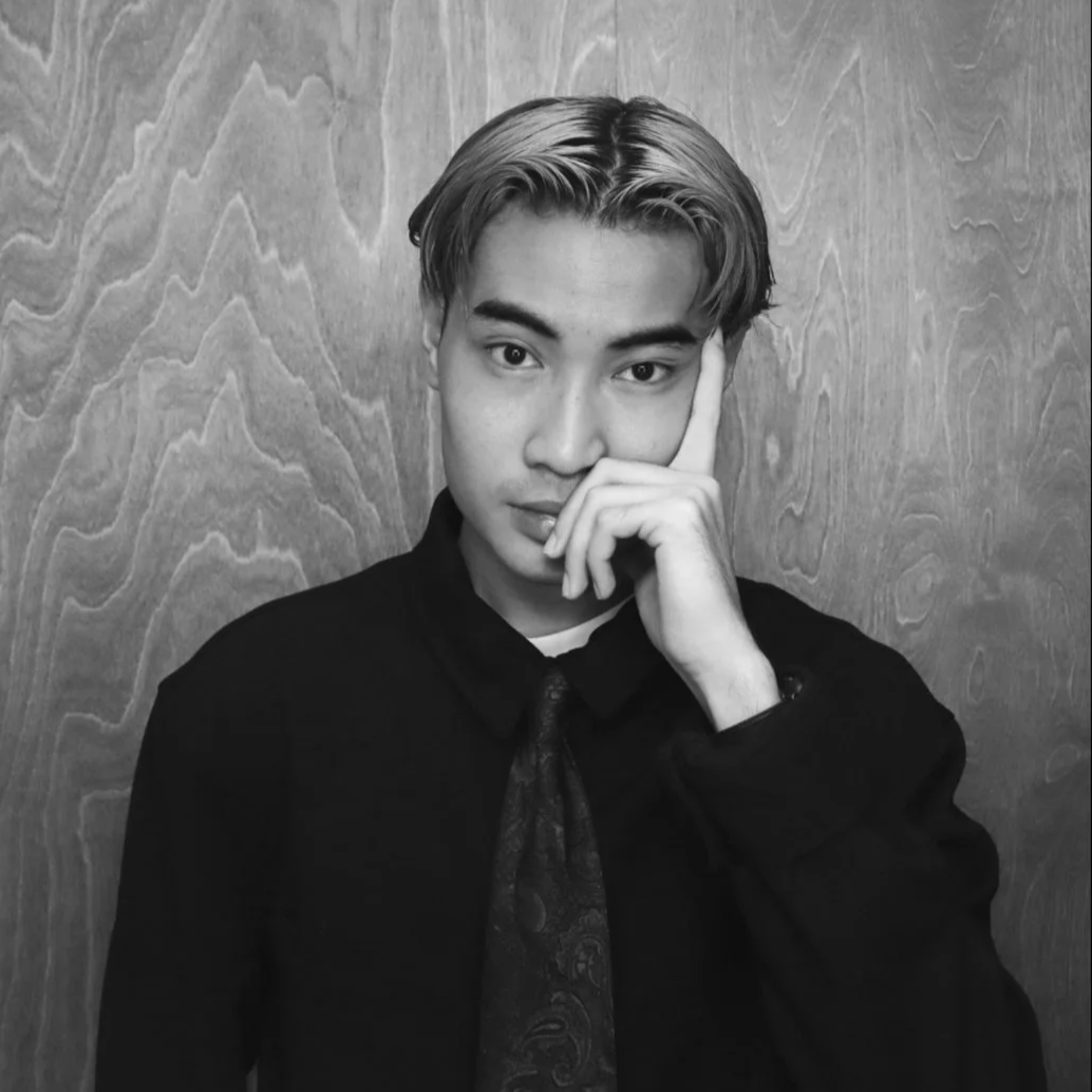 Black and white portrait of a young man with styled hair, wearing a dark shirt and tie, standing against a wooden background, with his right hand resting on his face.