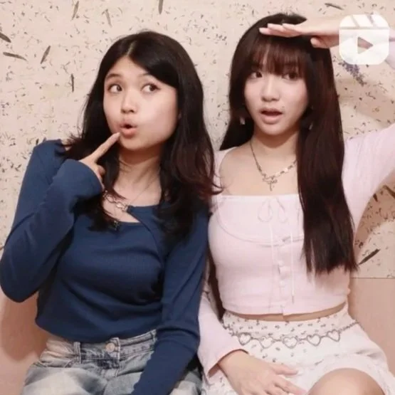 Two young women sitting against a beige wall, one wearing a blue top and the other a pink top, both making playful gestures with their faces.