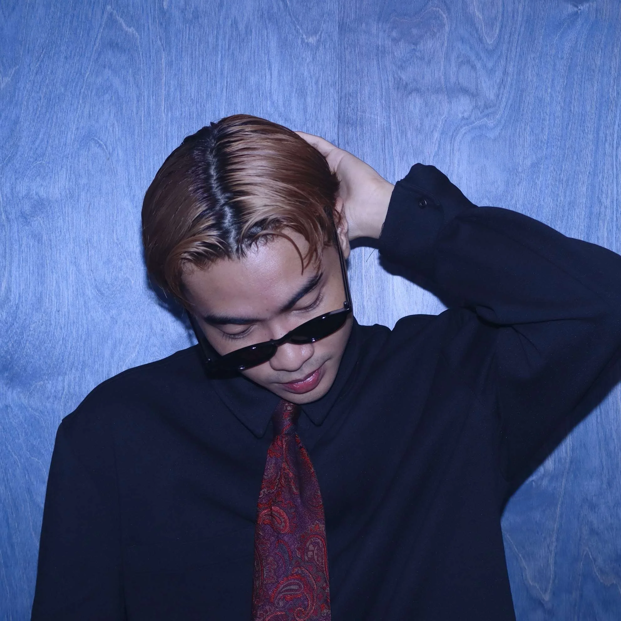 A person with brown hair wearing black sunglasses, a black shirt, and a red patterned tie, resting their head on a blue surface.