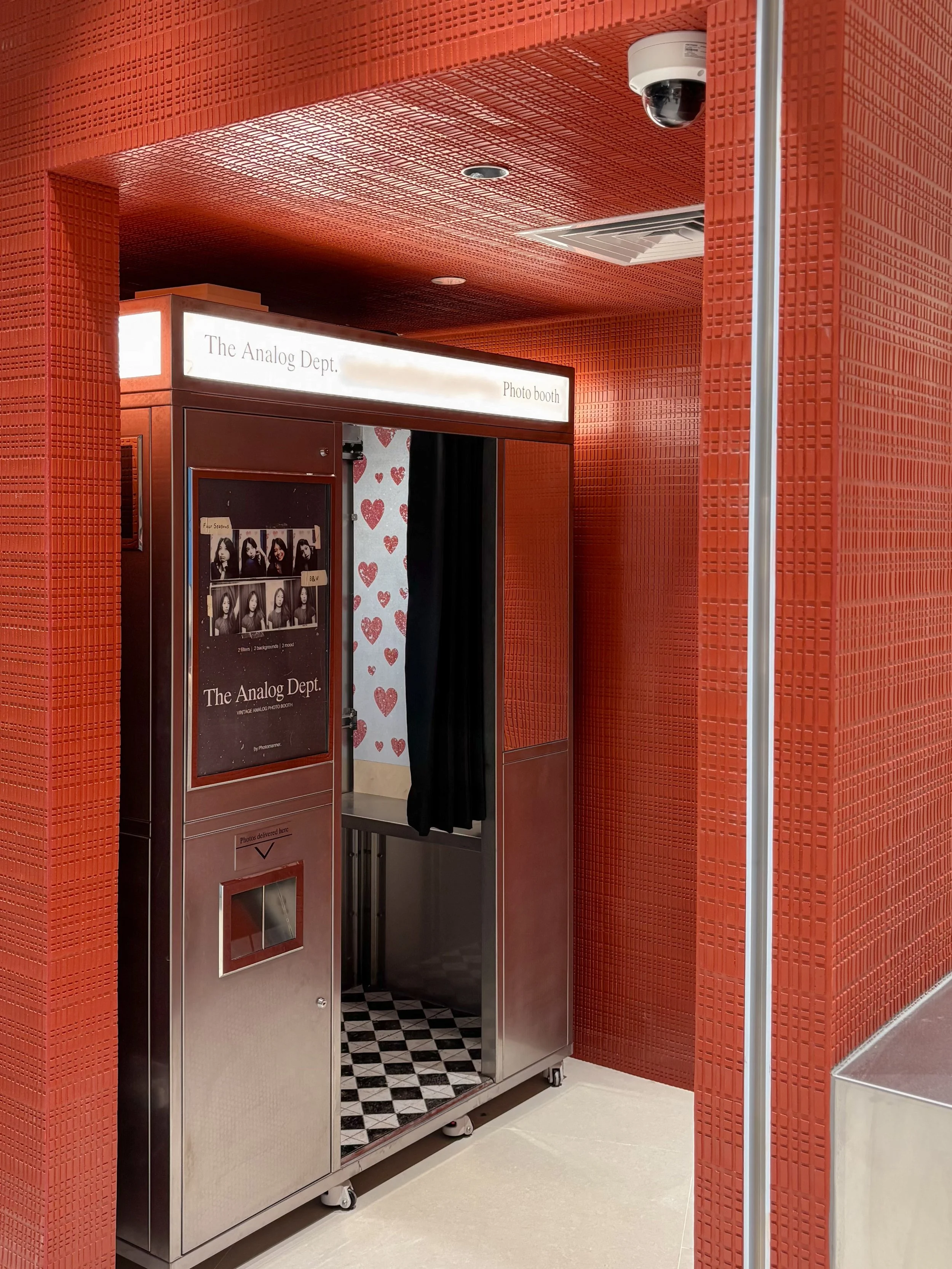 Photo booth with a sign that says "The Analog Dept." and a display case showing photo strips, inside a red-tiled room with a security camera and a pole.