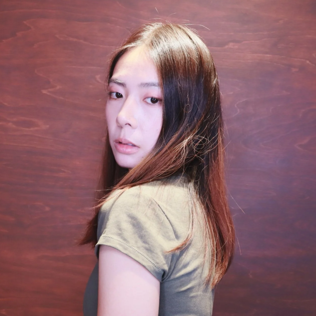 A young woman with long brown hair turned to the side, looking at the camera, against a wooden wall background.