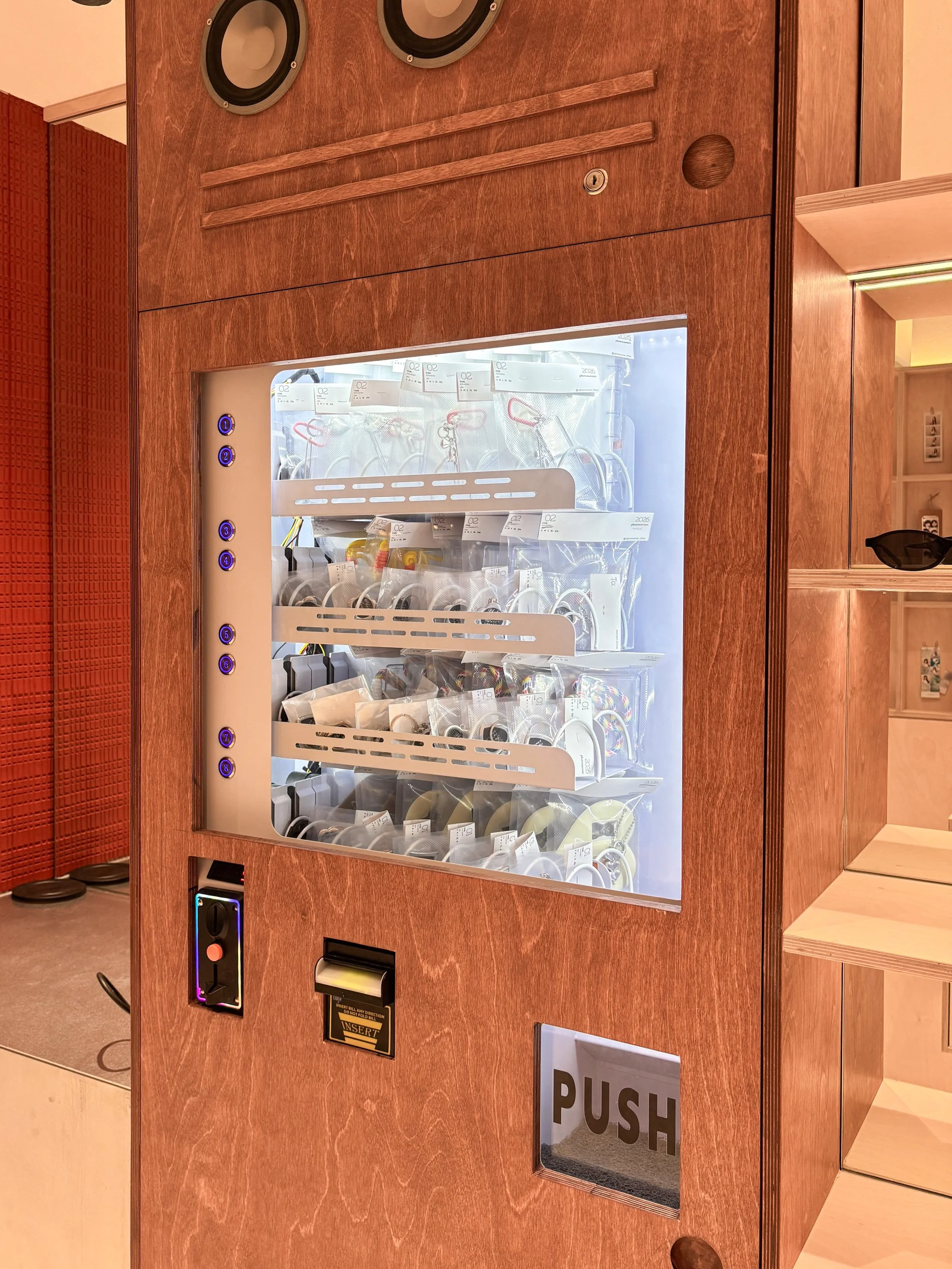 A wooden vending machine with a glass front displaying various snacks and small items inside. It has blue and purple buttons and a coin slot on the side. There is a