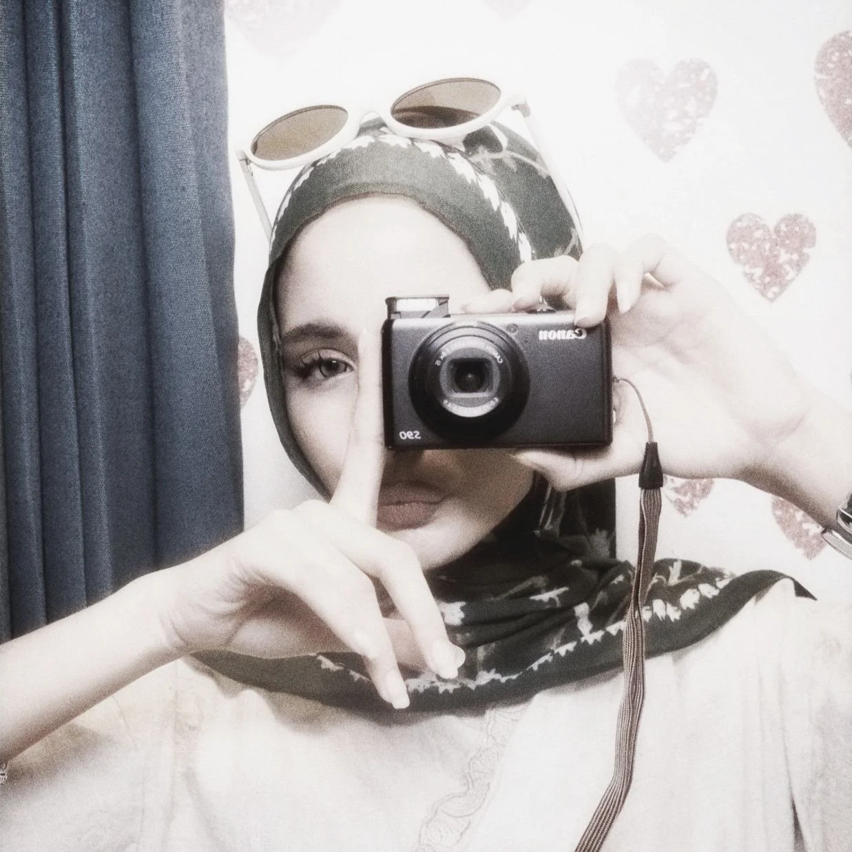 A woman taking a mirror selfie with a camera, wearing a headscarf and sunglasses on her head, with a wall decorated with heart patterns behind her.