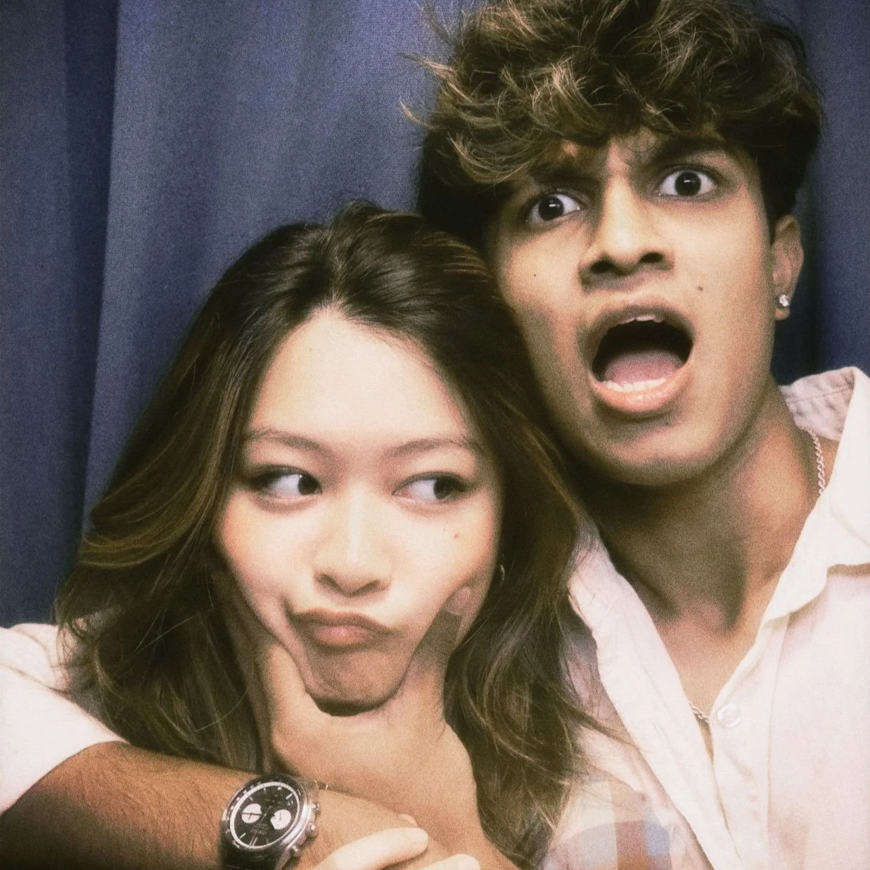 A woman and a man taking a selfie together. The woman is making a playful face by pursing her lips and resting her chin on her hand. The man has a surprised or shocked expression with his mouth open. They are close to each other, with the woman resting her head on the man's arm. The background appears to be a dark curtain.