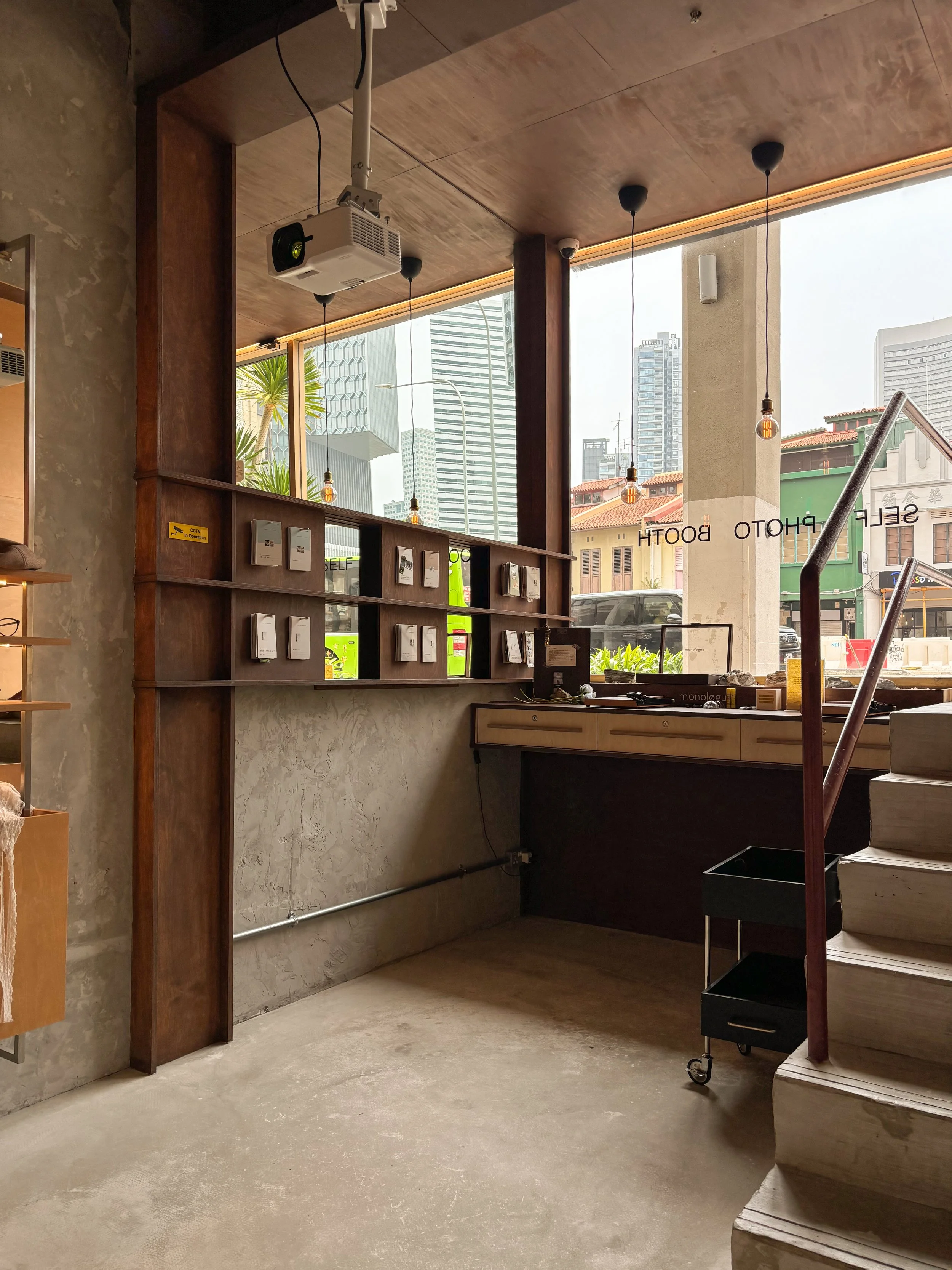 Interior of a modern store or cafe with large windows, a wooden ceiling, hanging light bulbs, and a display shelf with various pamphlets or menus. Outside city buildings are visible through the windows.