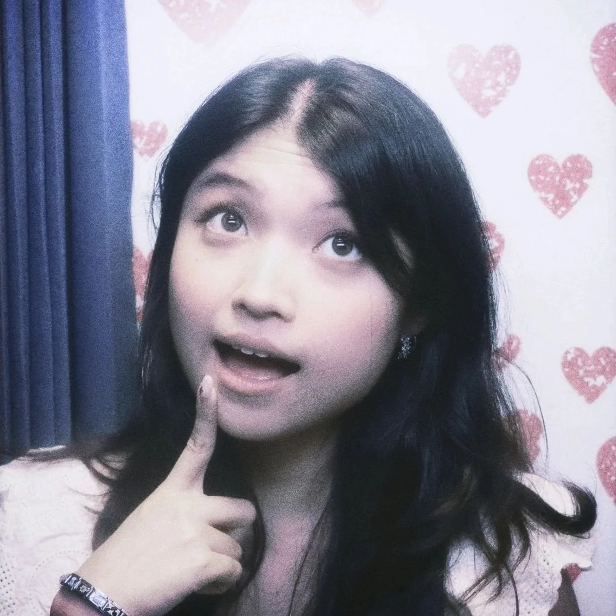 A young woman smiling slightly, in front of an Analog Photobooth backdrop with pink heart patterns.