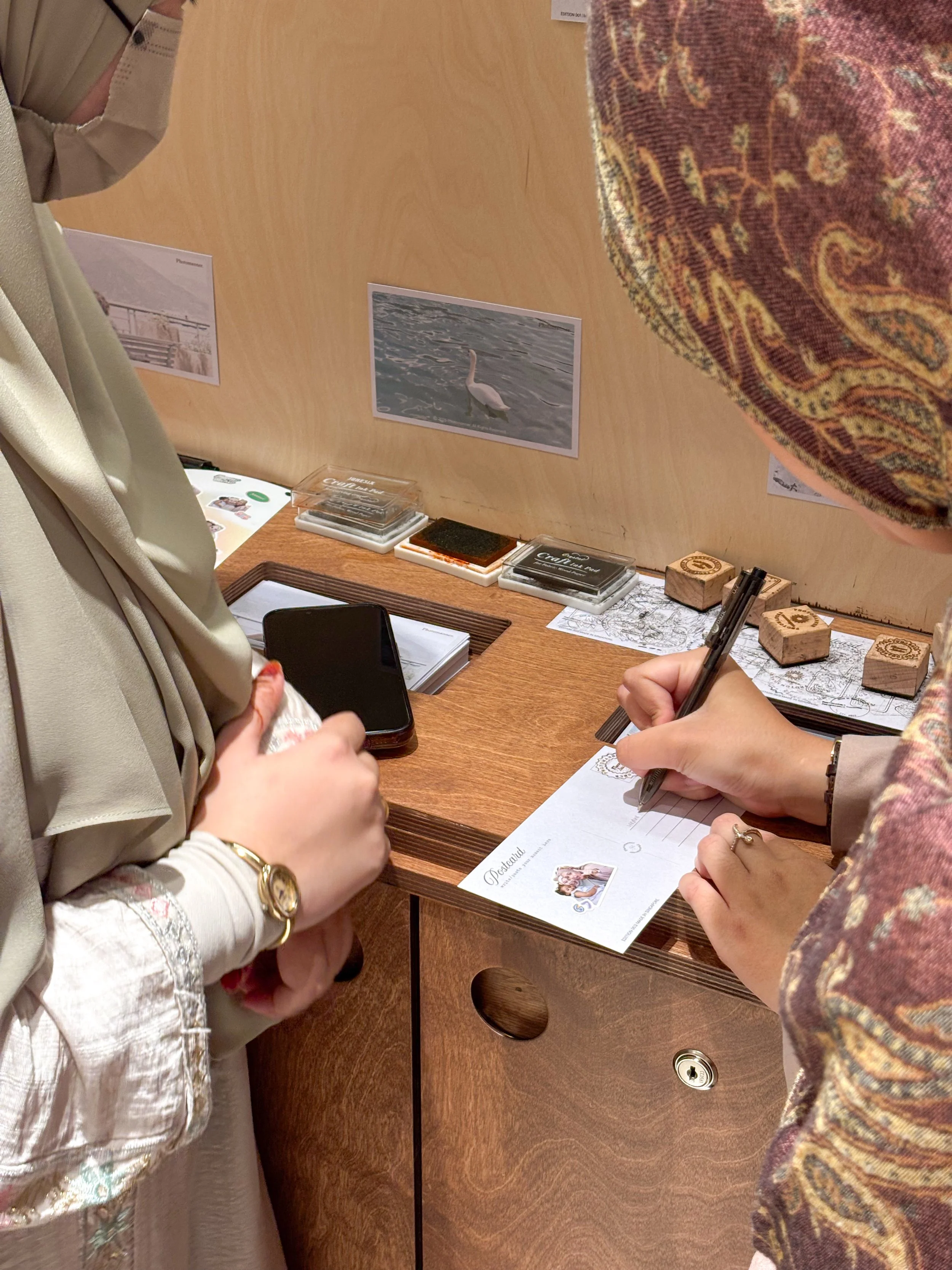 Two women are signing a guestbook at a desk. One is wearing a beige shirt with floral embroidery, the other has a patterned headscarf. There are prints of a swan, a bridge, and a ship on the wall behind them. The desk has stamps, a smartphone, and a brochure.