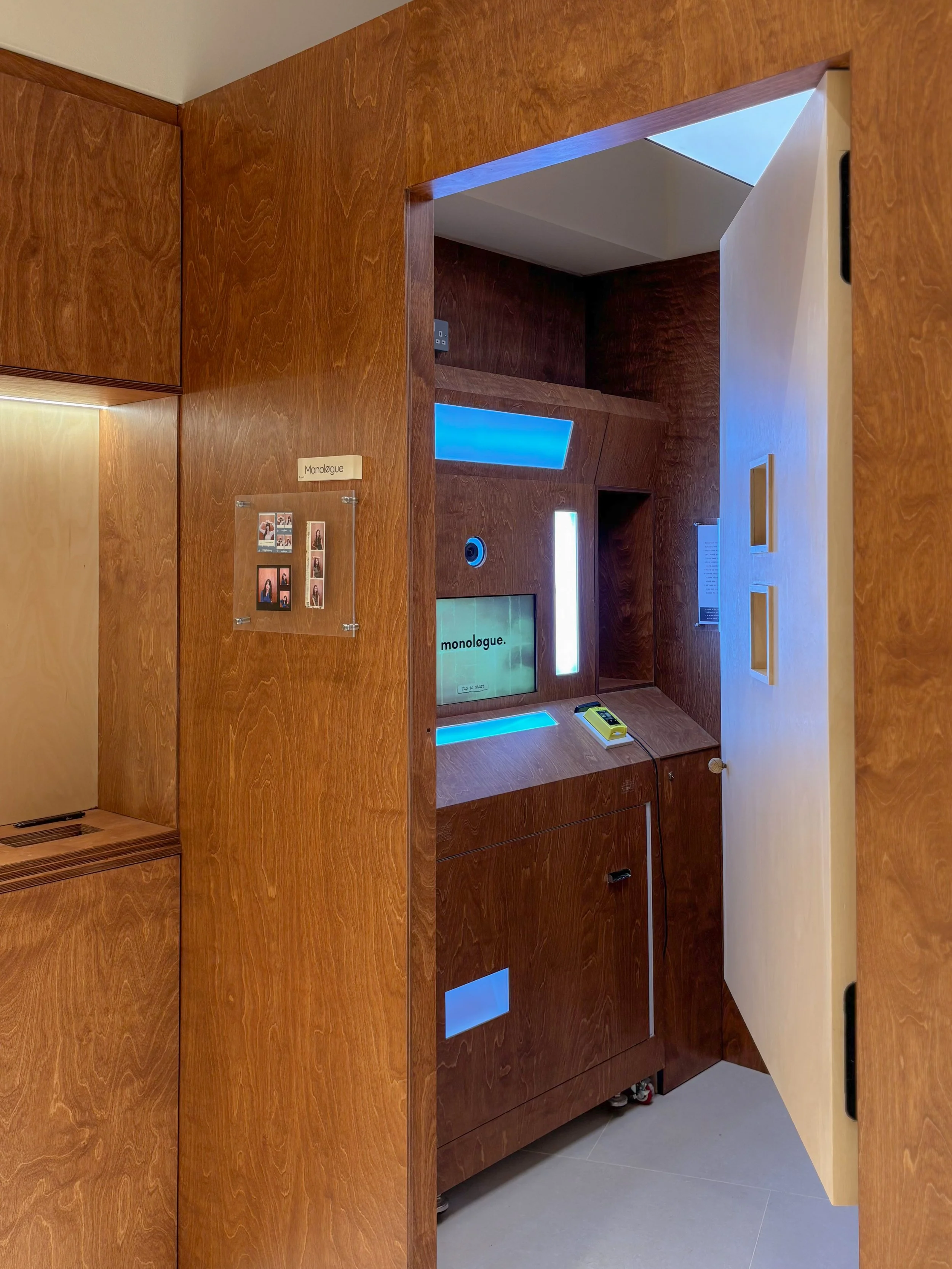 Interior of a room with wood-paneled walls and a monologue station with a digital screen, a card reader, and a yellow device, under a ceiling with blue lighting accents.