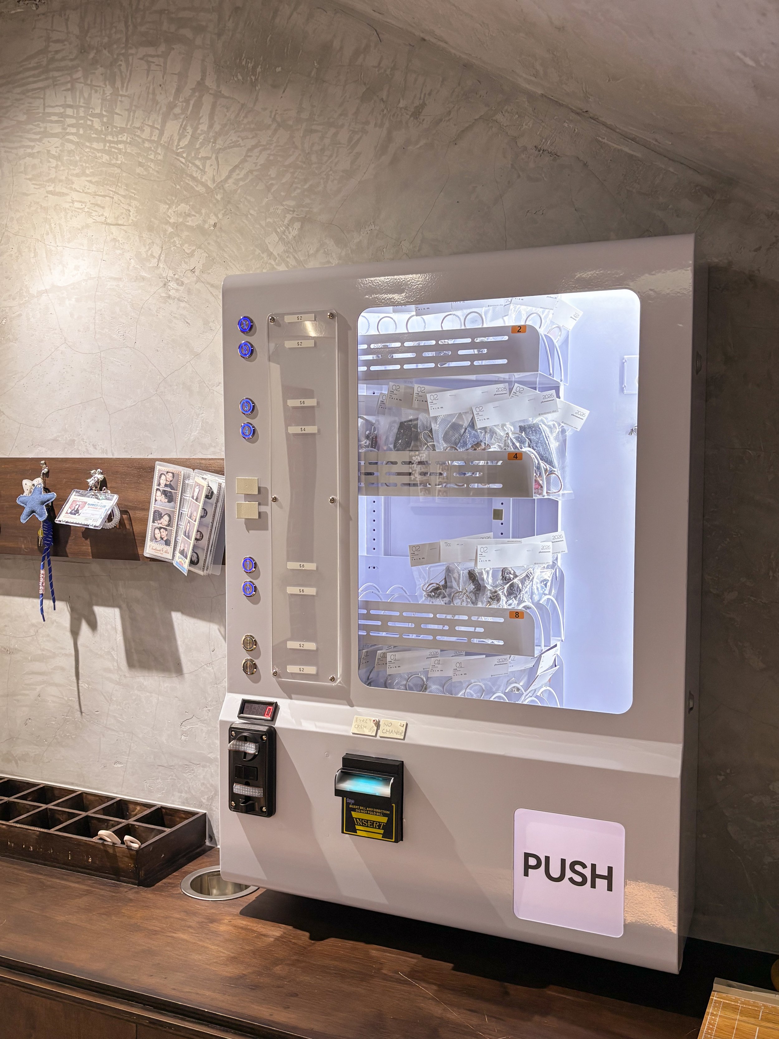 A white vending machine with a glass front filled with various items inside, with blue illuminated buttons on the left side, labeled with prices. There is a yellow and black card reader at the bottom, and a white sign with black text that says 'PUSH'. The vending machine is on a wooden surface, and the background features a textured beige wall and a small table with photo frames and decorative items.