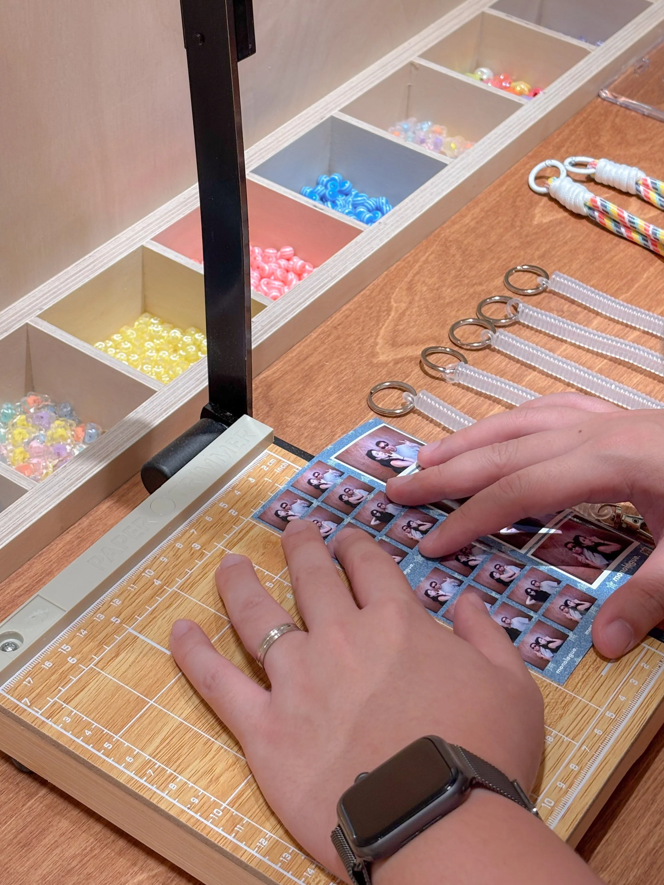 A person is arranging small photo booth pictures on a cutting mat on a wooden table. The person wears a black wristwatch and a silver ring.